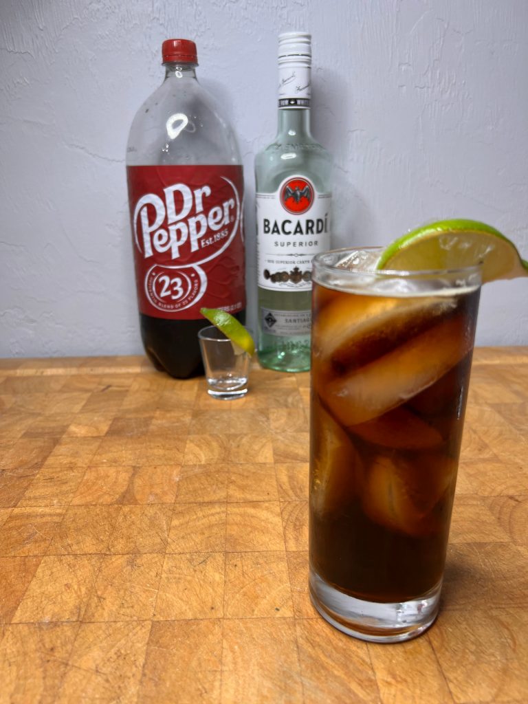 close up of dr pepper & rum with liquor bottles visible in the background