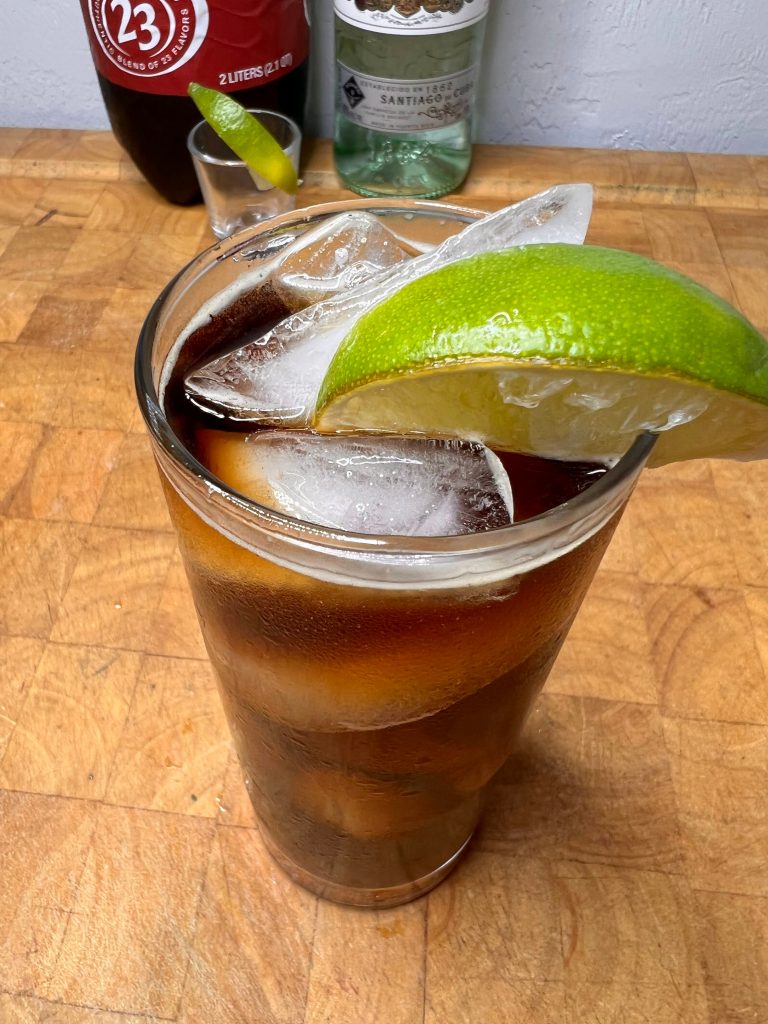 close up of dr pepper & rum with liquor bottles visible in the background