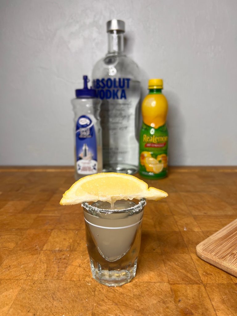 close up of lemon drop shot with liquor bottles visible in the background
