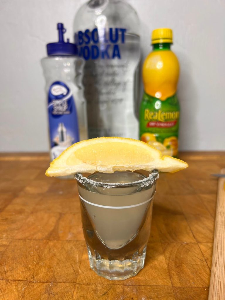 close up of lemon drop shot with liquor bottles visible in the background