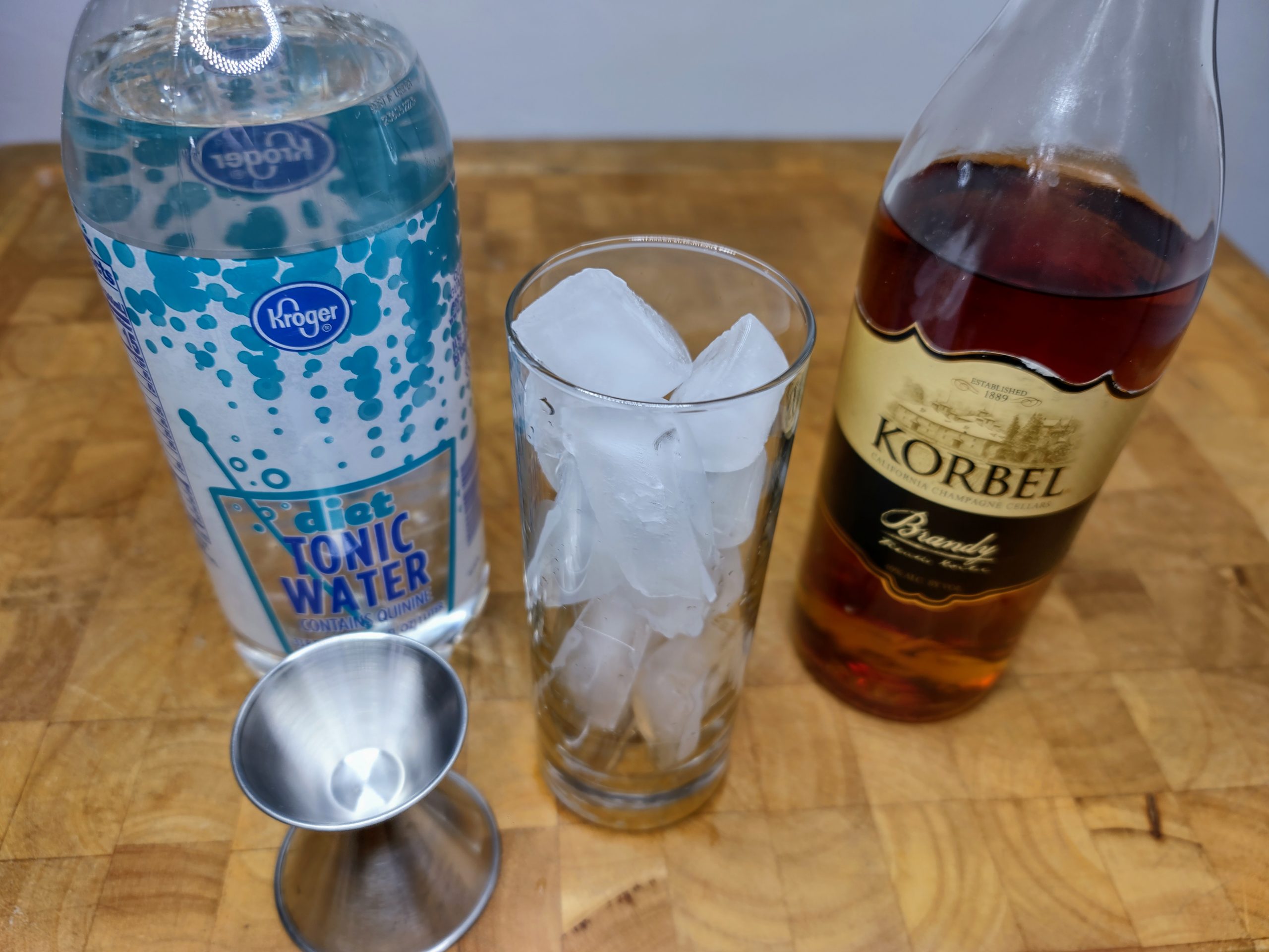 jigger, highball glass filled with ice, bottle of brandy and bottle of tonic water sitting on a wooden table