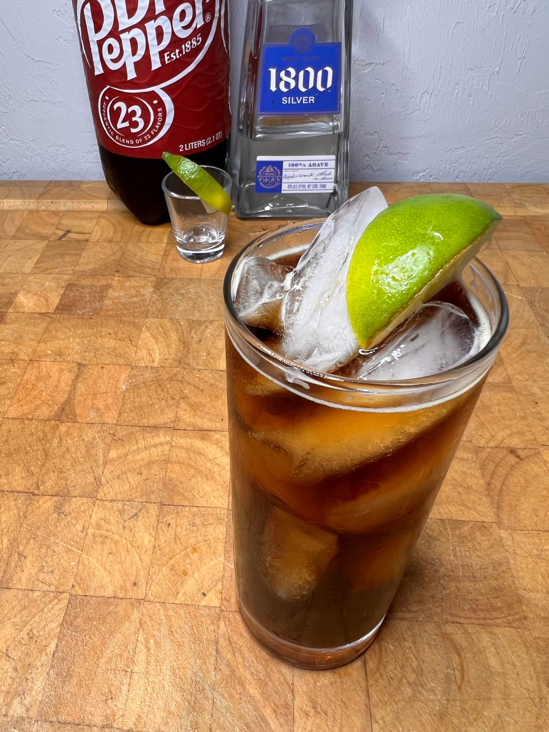 tequila and dr pepper with ingredients behind the drink on a wooden table