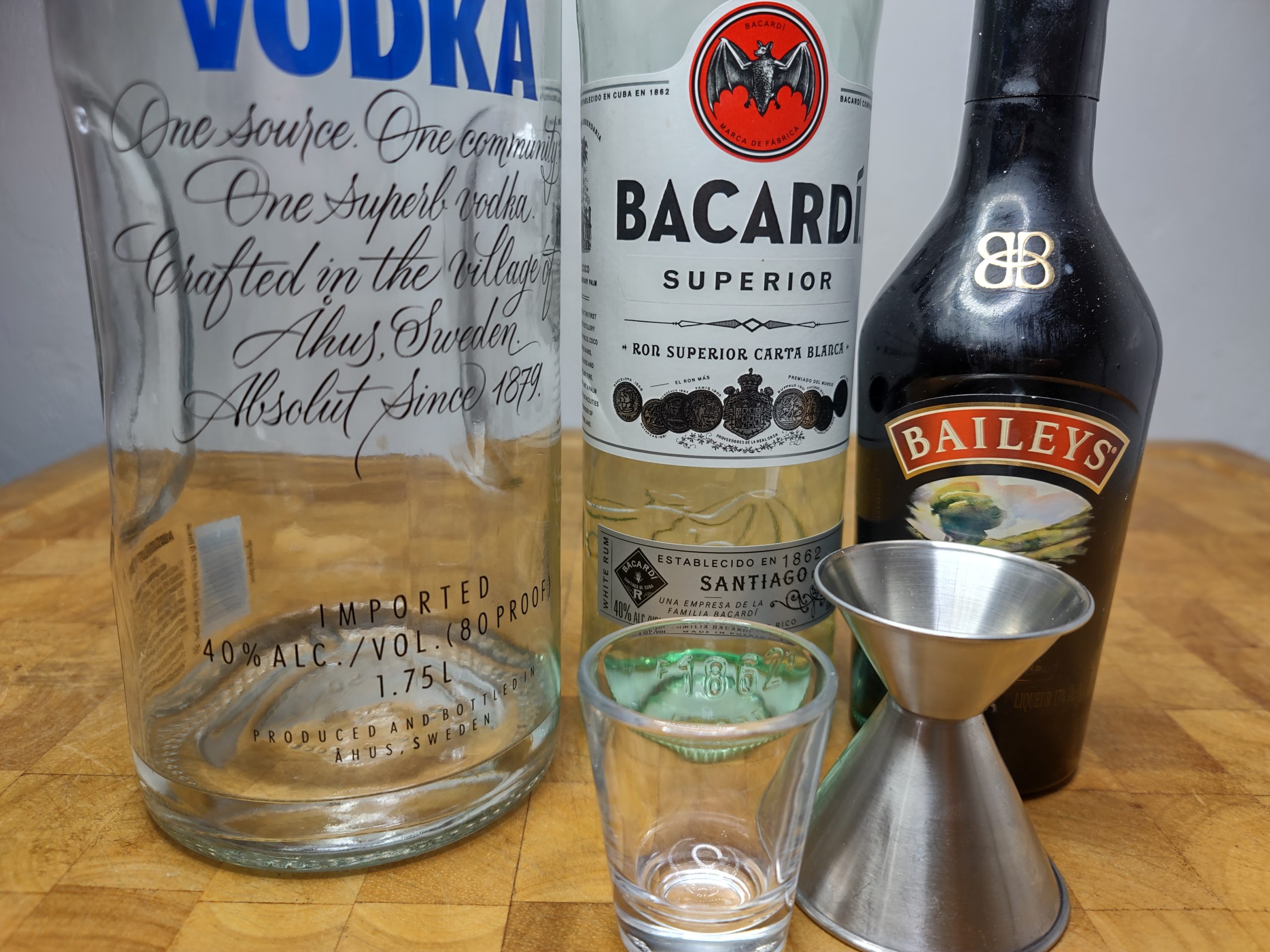 empty shot glass next to a jigger, bottle of vodka, white rum and irish cream