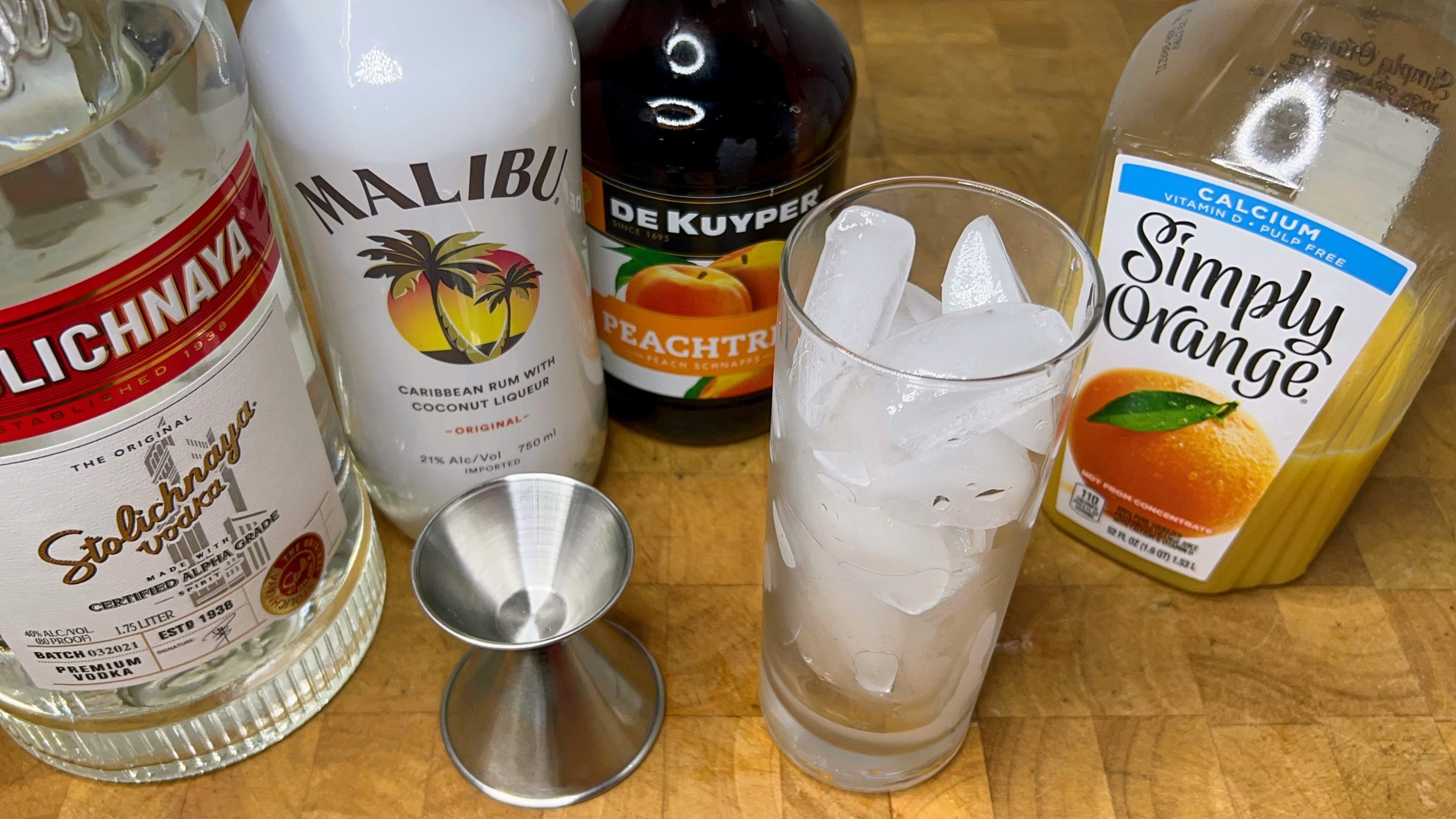 highball glass with ice next to jigger, orange juice, peach schnapps, vodka and coconut rum