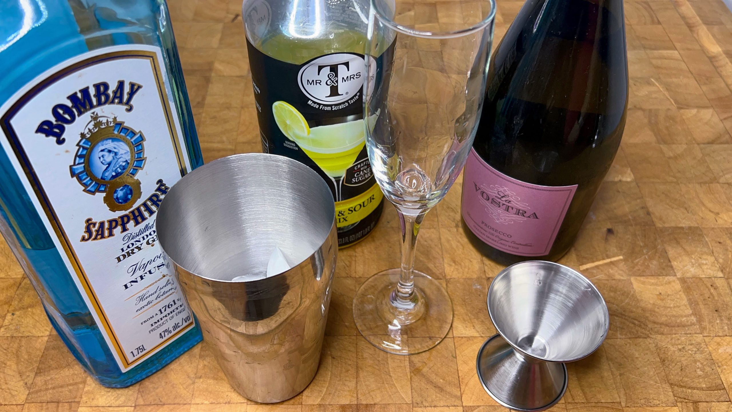 empty champagne flute next to jigger, cocktail shaker, simple syrup, gin and champagne