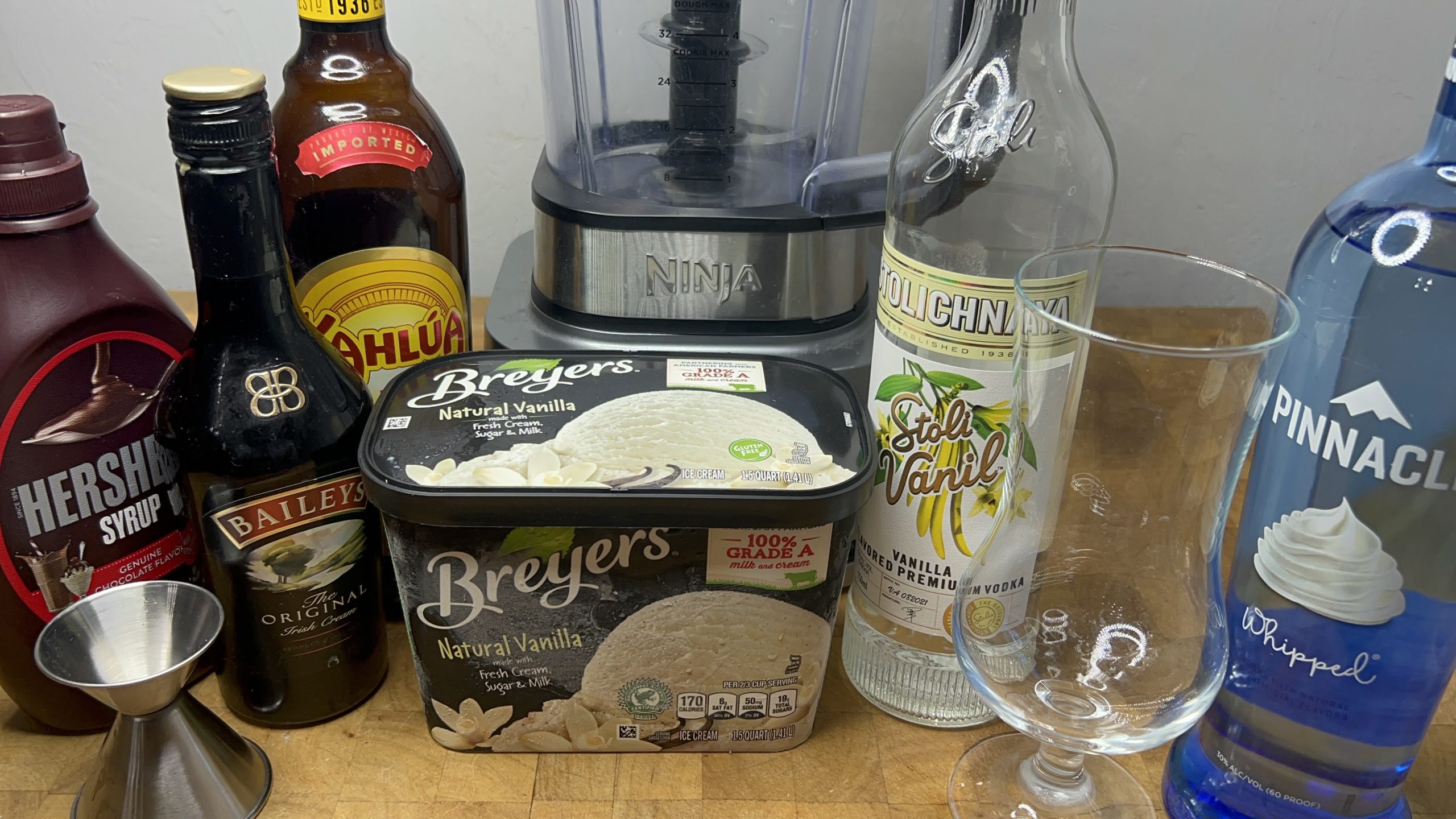 empty hurricane glass next to jigger, blender and all the ingredients used for a frozen mudslide