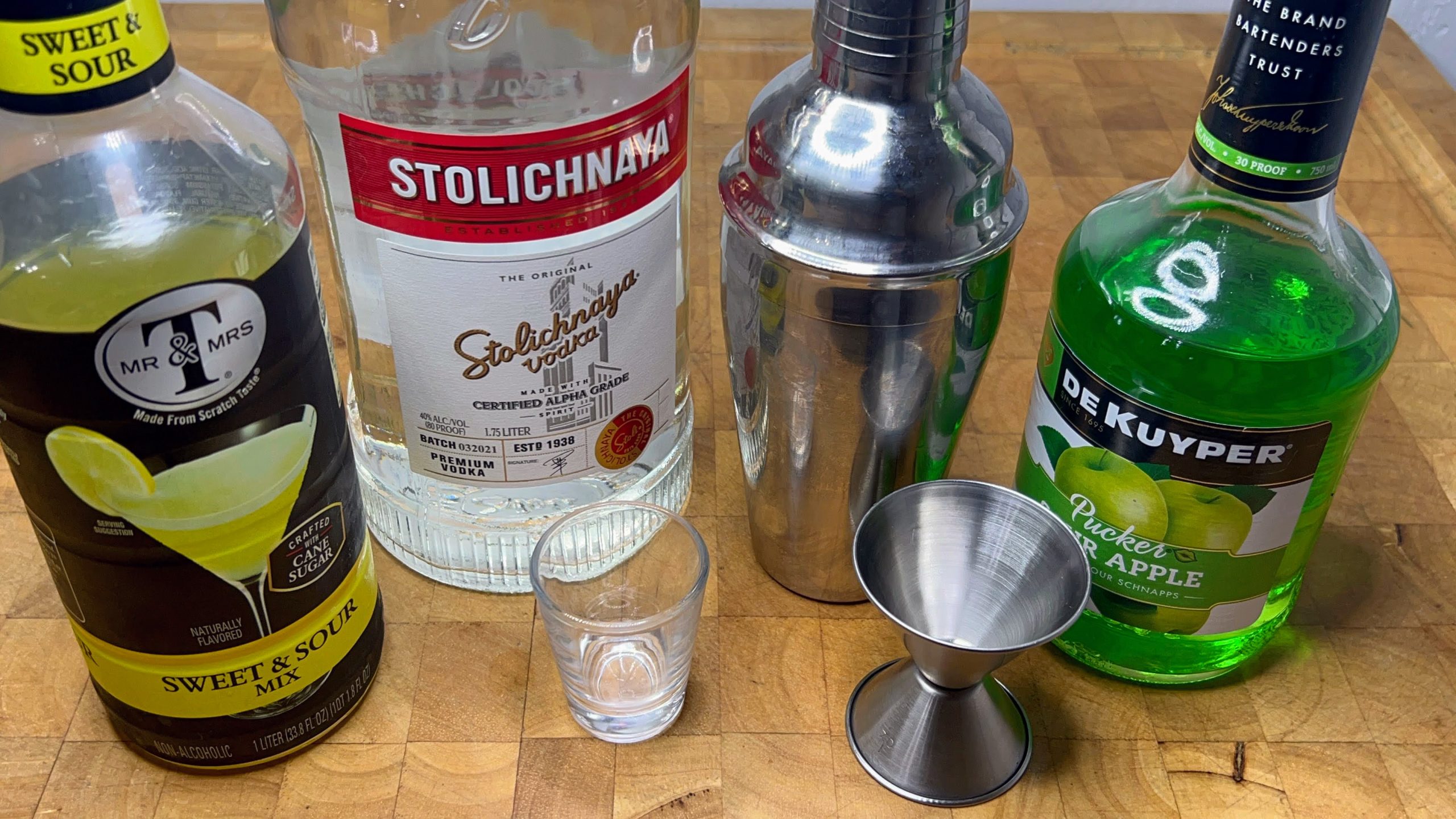 shot glass next to jigger, sour apple schnapps, sweet and sour mix, vodka and a cocktail shaker