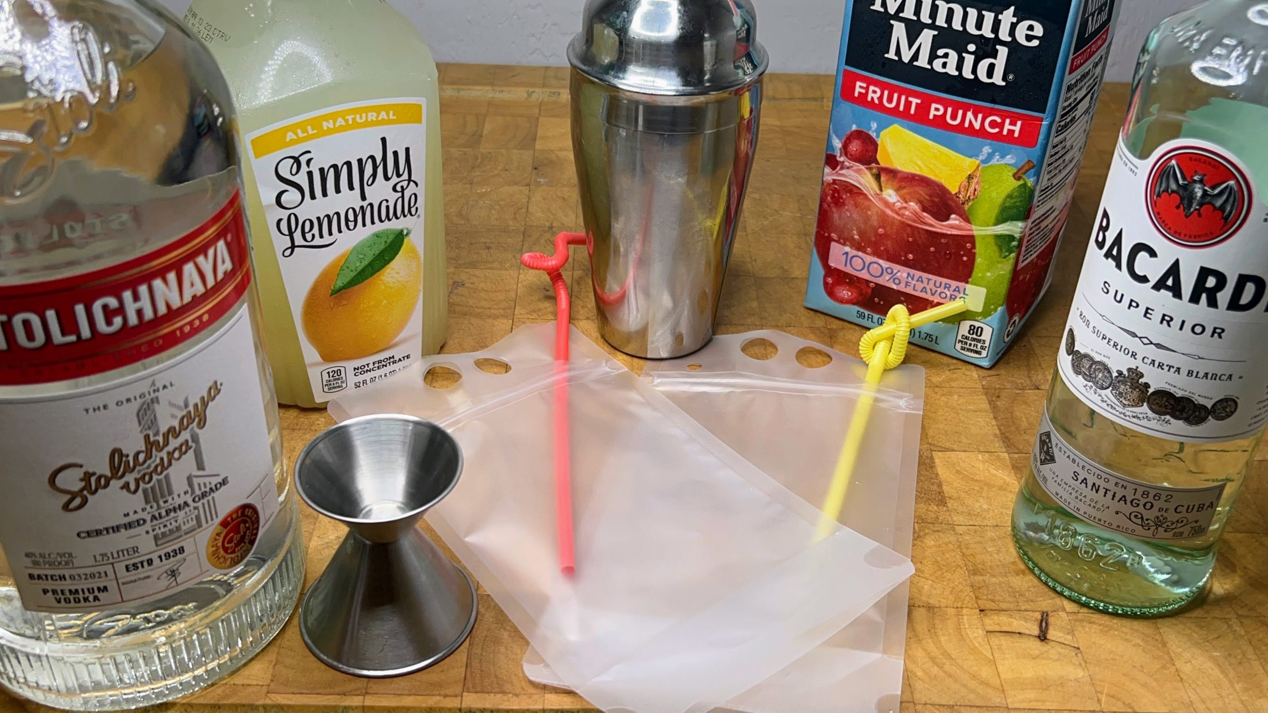 two plastic drink pouches with straws next to a jigger, cocktail shaker and bottles of vodka, white rum, lemonade and fruit punch
