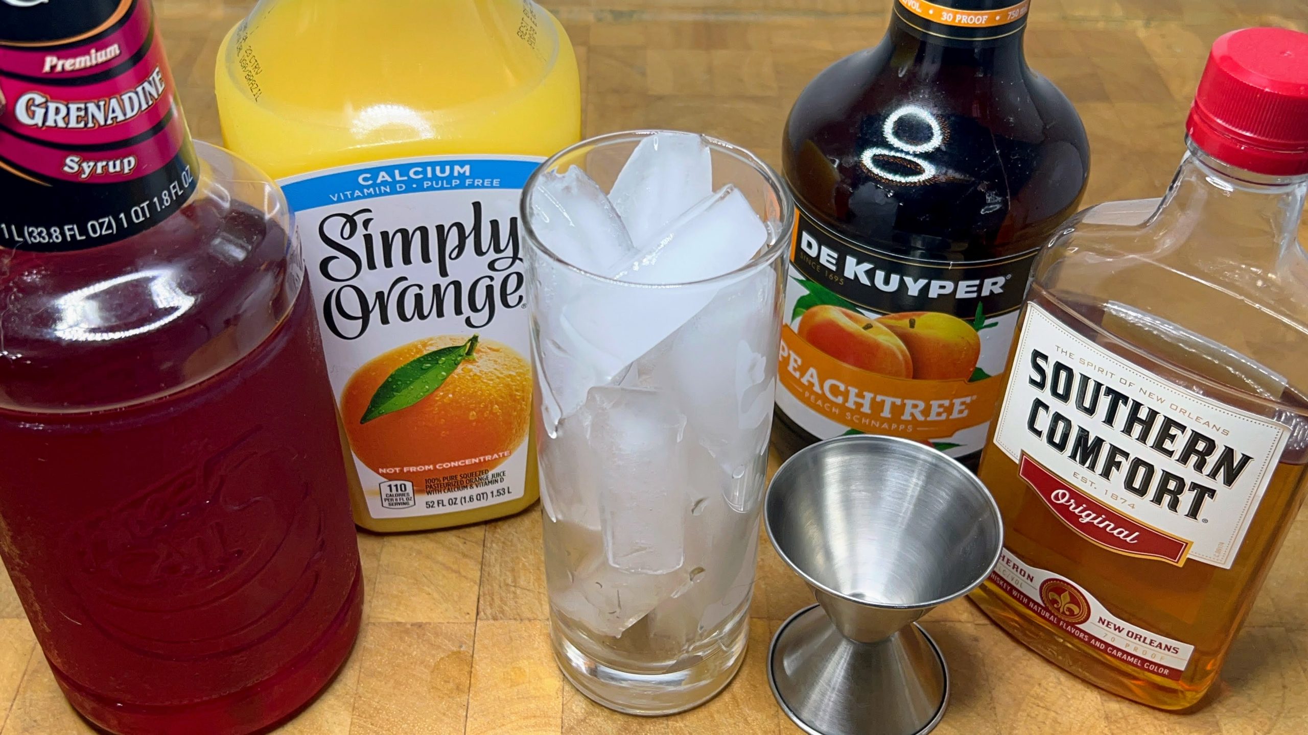 highball glass filled with ice next to jigger, and bottles of grenadine, OJ, peach schnapps and southern comfort