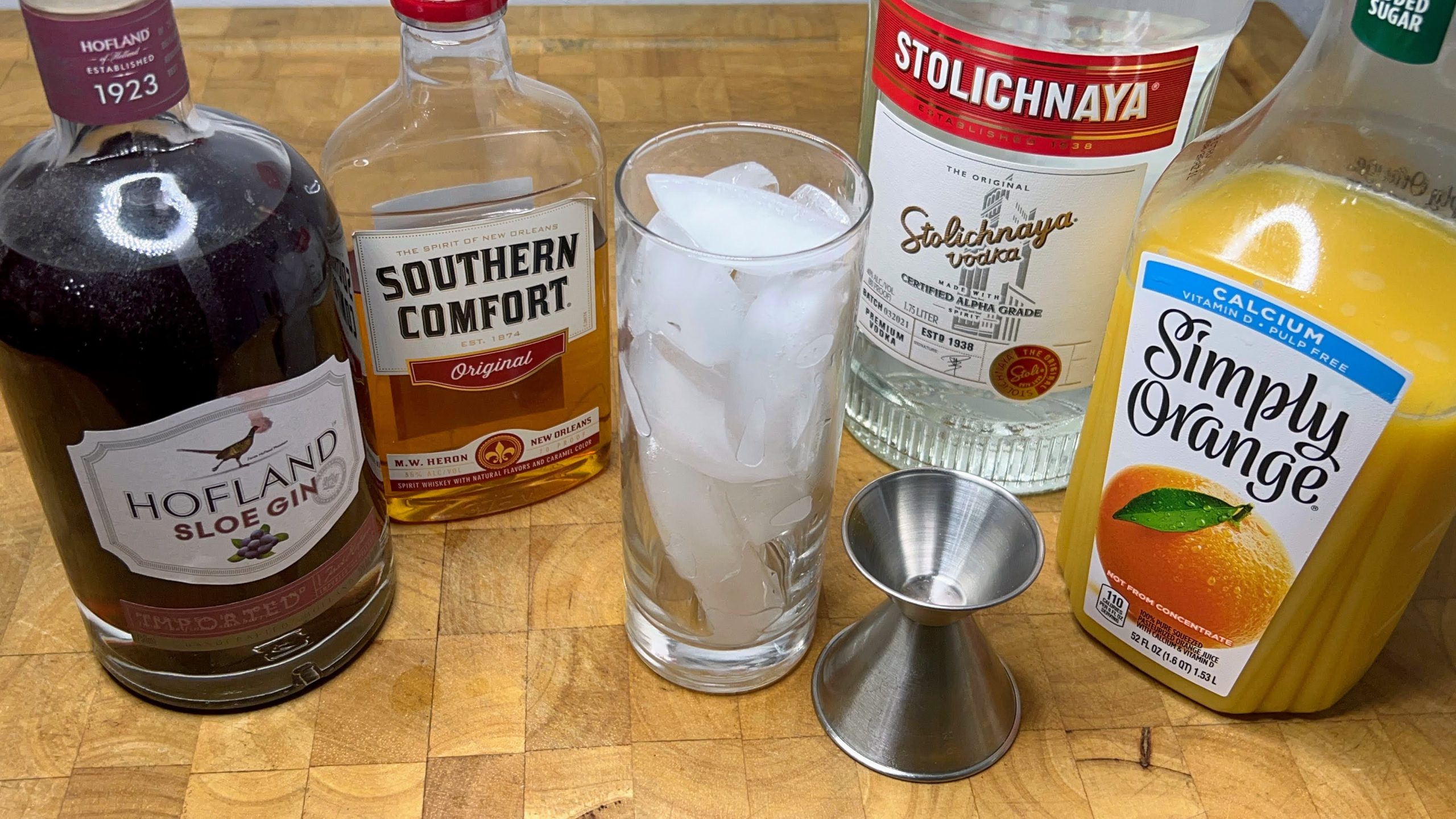 highball glass filled with ice next to a jigger and bottles of sloe gin, vodka, OJ and southern comfort
