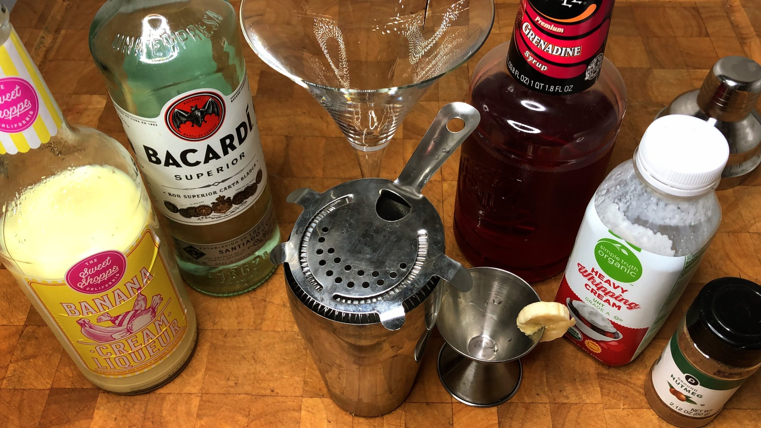 empty martini glass next to cocktail shaker, jigger with a slice of banana, banana liqueur, white rum, grenadine, nutmeg and heavy cream
