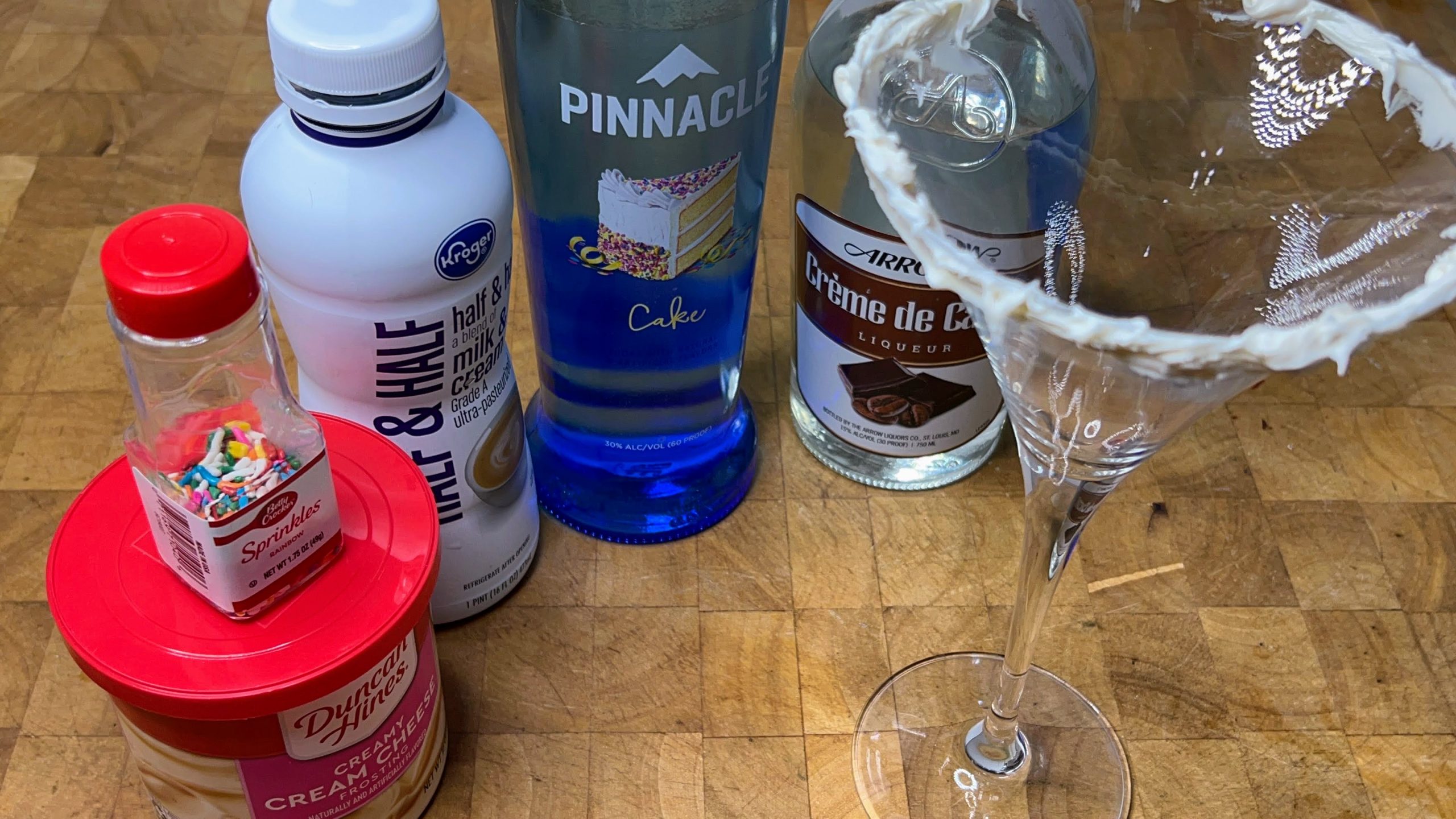 martini glass with frosting on the rim next to bottles of frosting, sprinkles, half and half, cake vodka and white cream de cacao