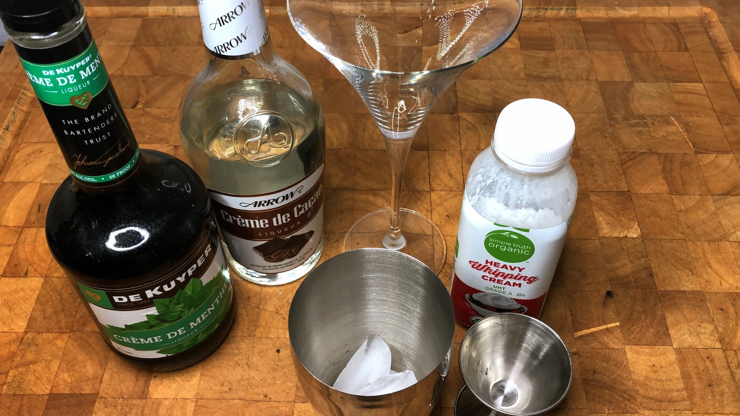 empty martini glass next to shaker, jigger, heavy cream, creme de cacaao and creme de menthe