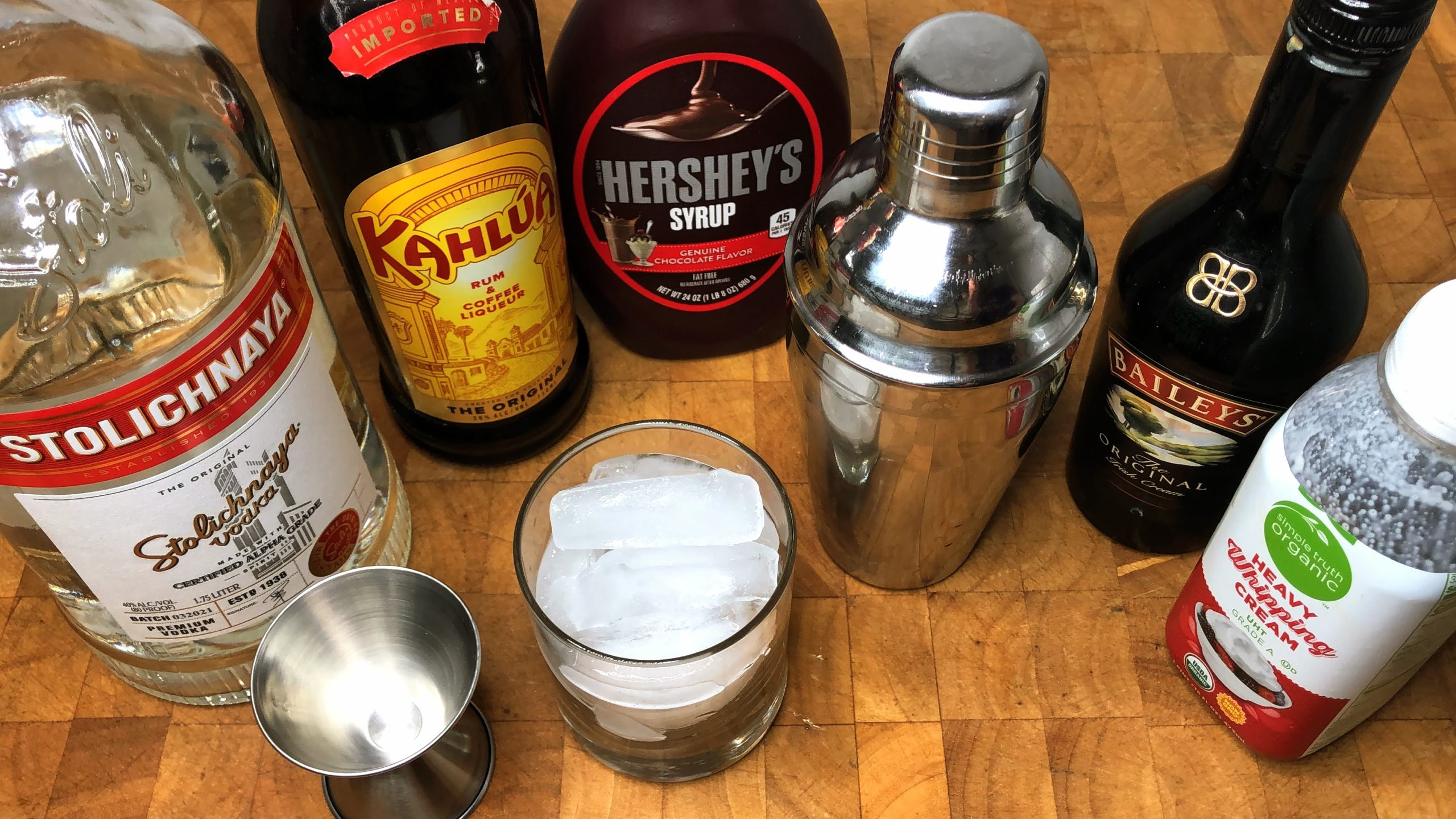 rocks glass filled with ice next to jigger, cocktail shaker, vodka, kahlua, chocolate syrup, irish cream and heavy cream