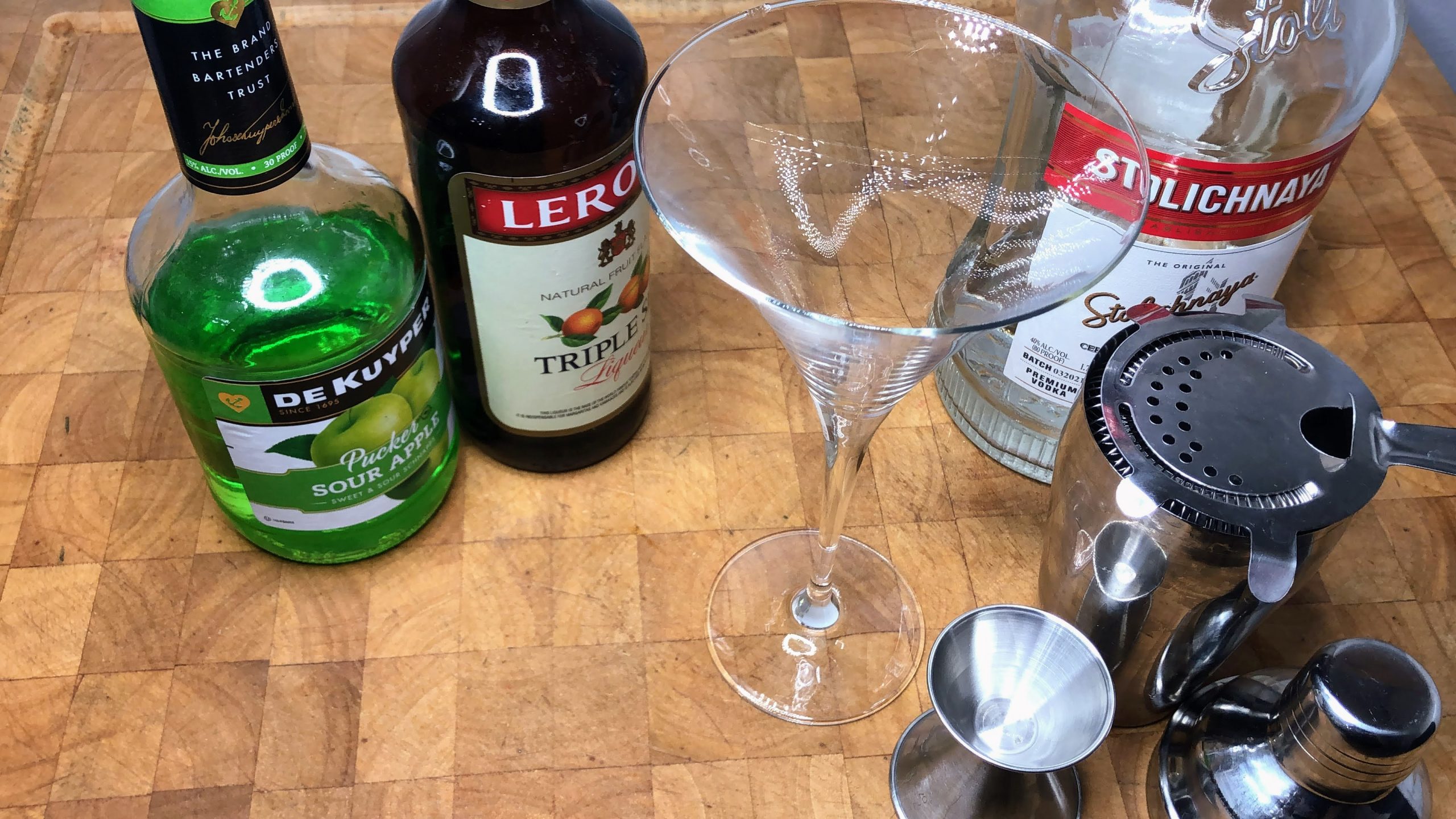 martini glass next to cocktail shaker, jigger, vodka, triple sec and sour apple pucker
