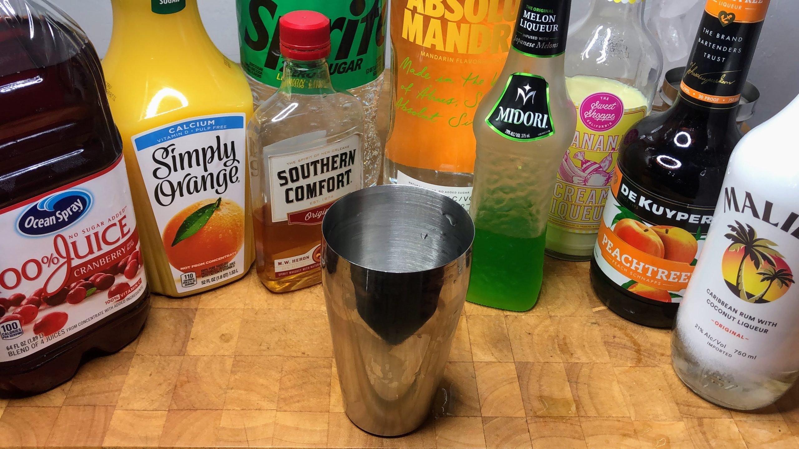 cocktail shaker in front of bottles of Soco, OJ, midori, vodka, sprite, cranberry juice, banana liqueur, coconut rum and peach schnapps