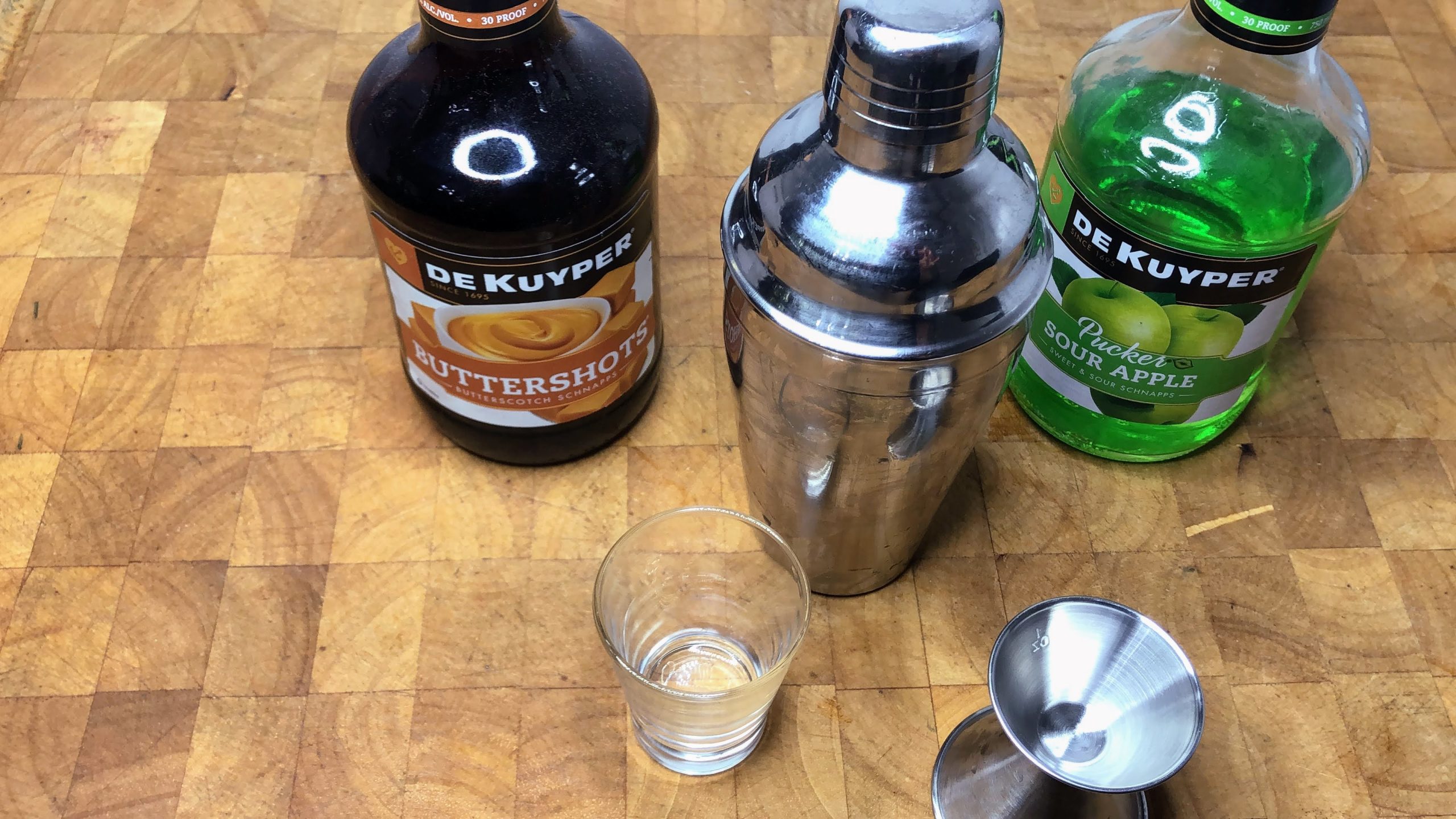 shot glass next to jigger, shaker and bottles of butterscotch schnapps and sour apple pucker