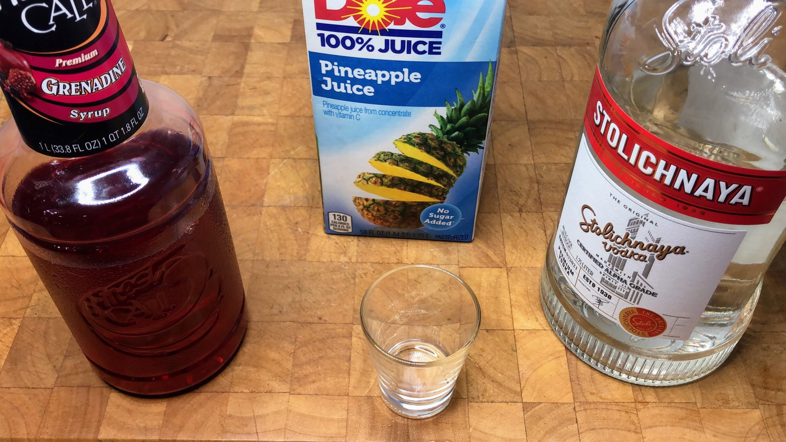 empty shot glass next to bottles of grenadine, bodka and pineapple juice
