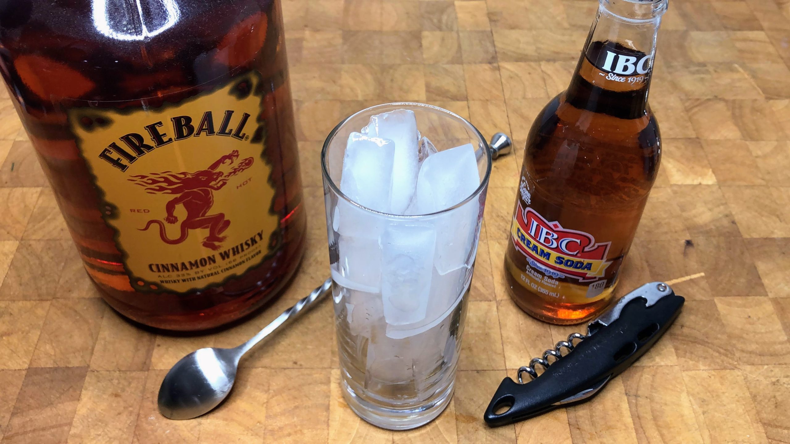 highball glass with ice next to bottle opener, bar spoon and bottles of fireball and cream soda