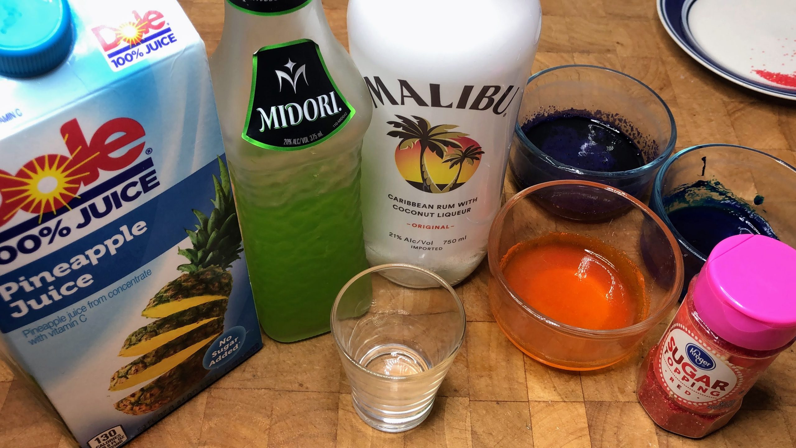 shot glass next to red sugar, orange sugar, purple sugar, blue sugar and bottles of malibu, midori and pineapple juice