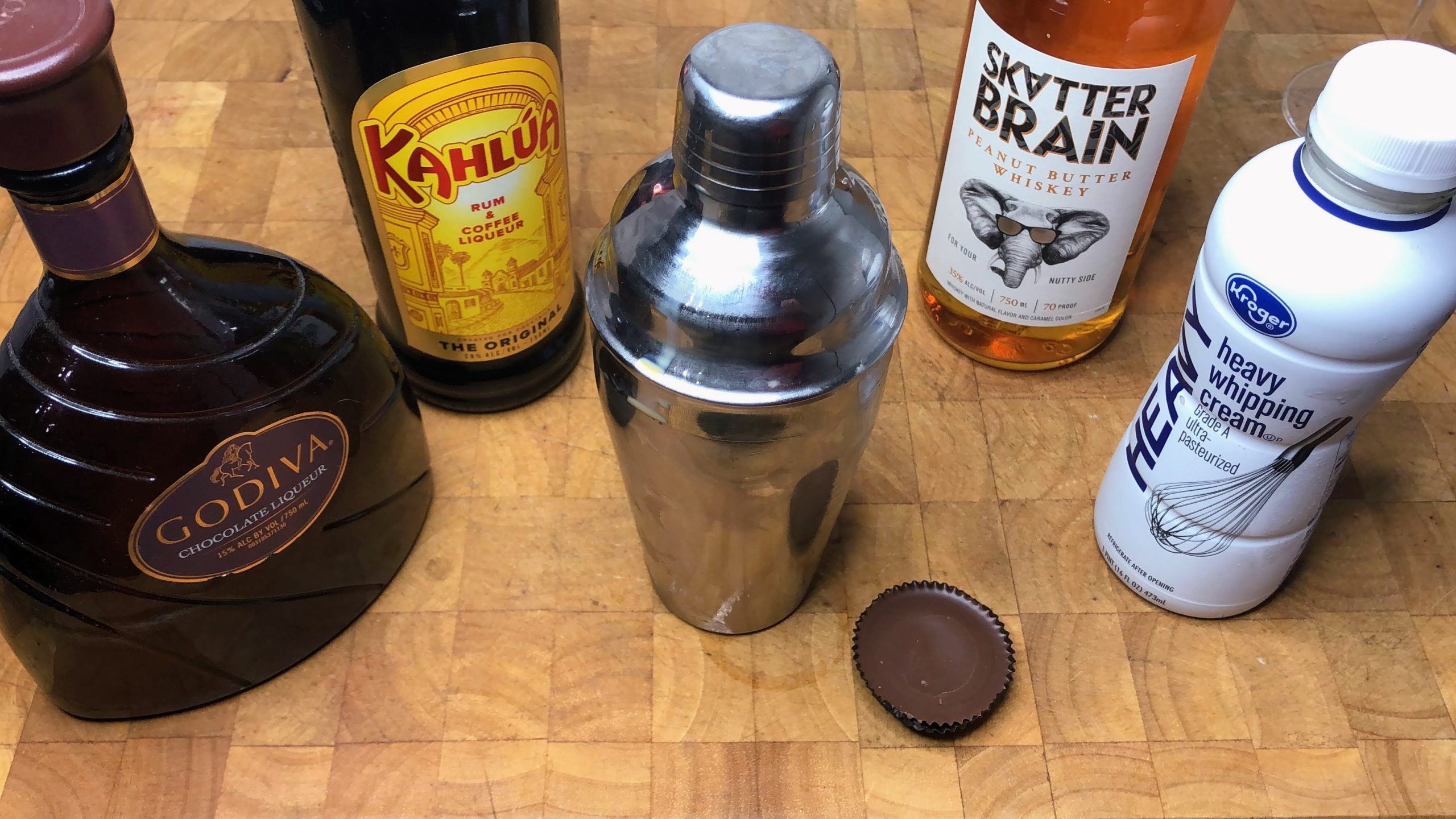 cocktail shaker next to peanut butter cup and bottles of godiva, kahlua, peanut butter whiskey and heavy cream