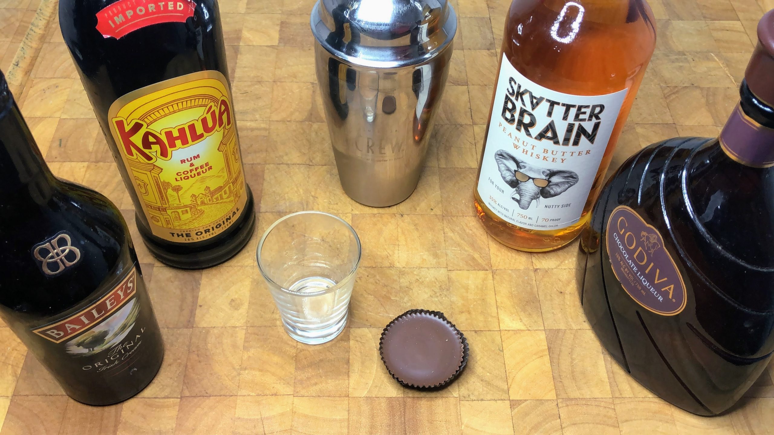 empty shot glass next to shaker, peanut butter cup and bottles of godiva, peanut butter whiskey, kahlua and irish cream