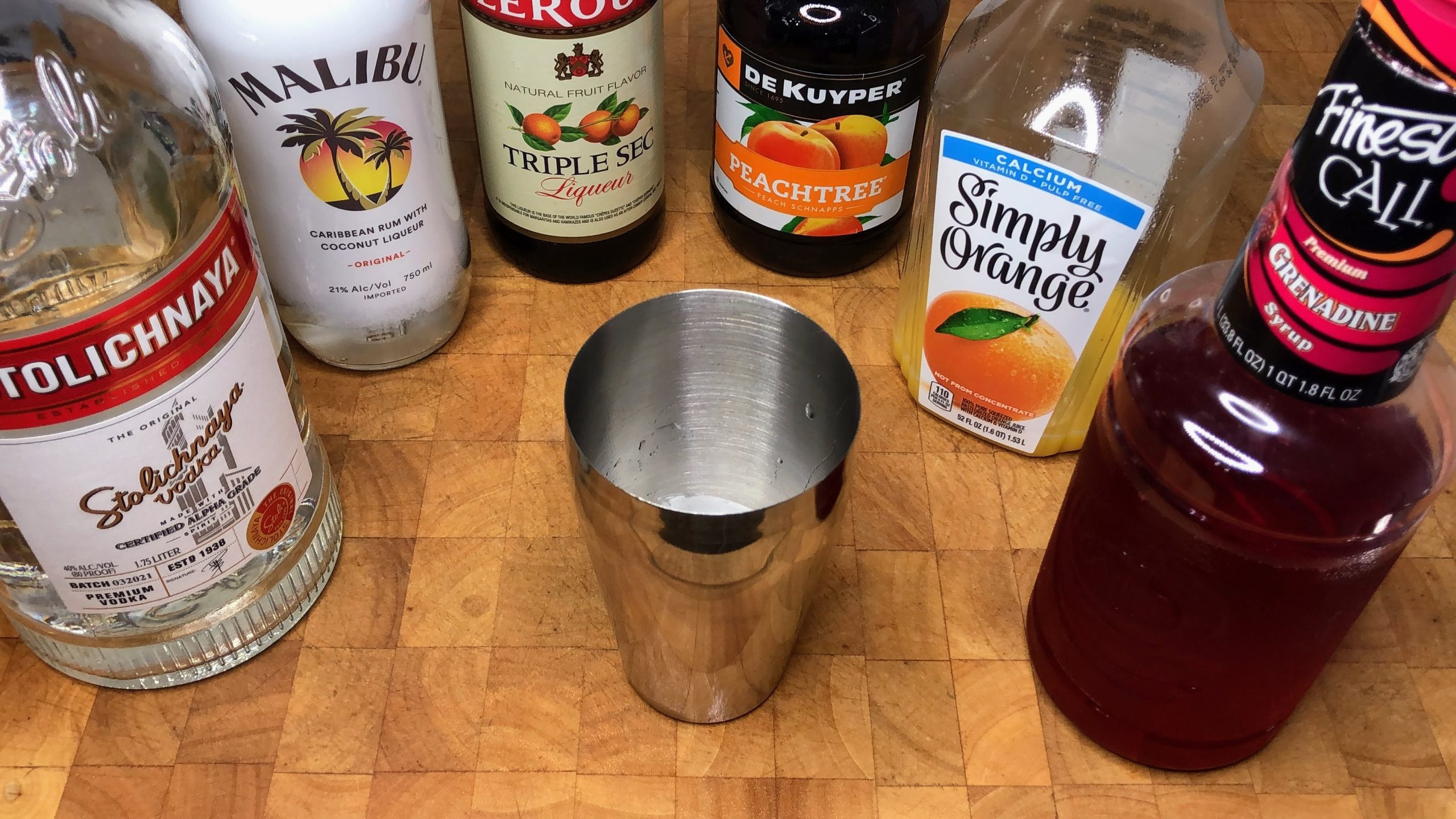 cocktail shaker next to bottles of grenadine, OJ, peach schnapps, triple sec, malibu and vodka