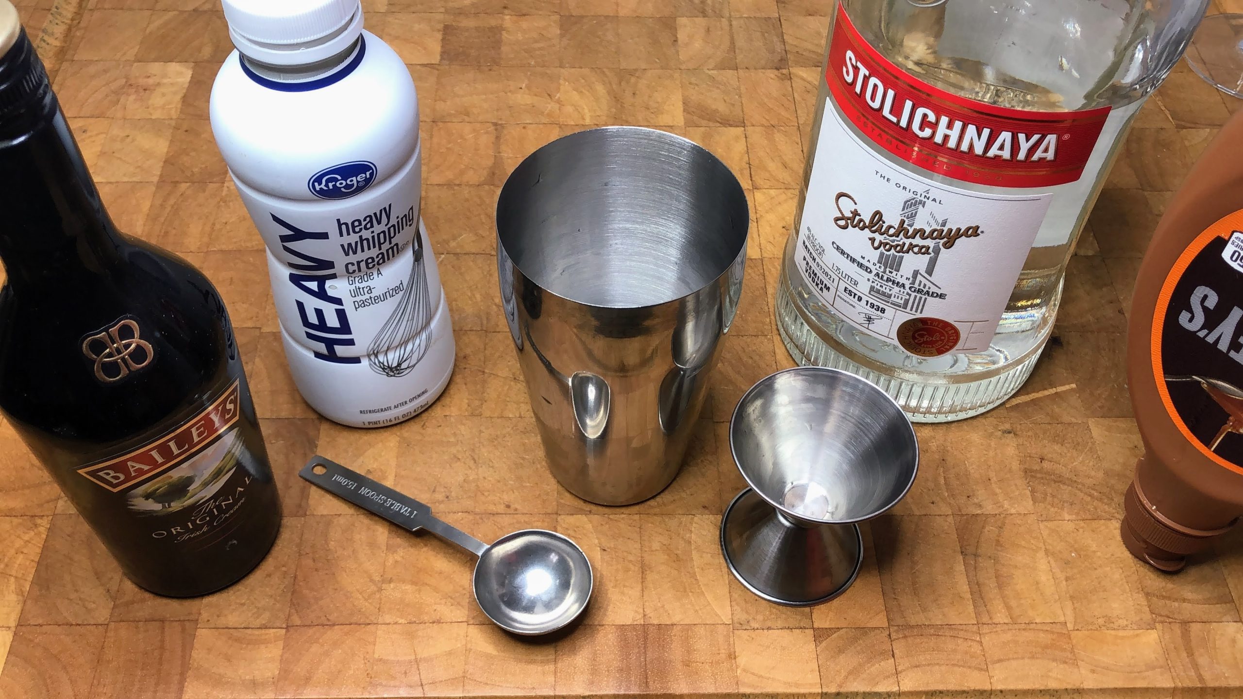 cocktail shaker next to teaspoon, jigger, caramel syrup and bottles of irish cream, heavy cream, vodka