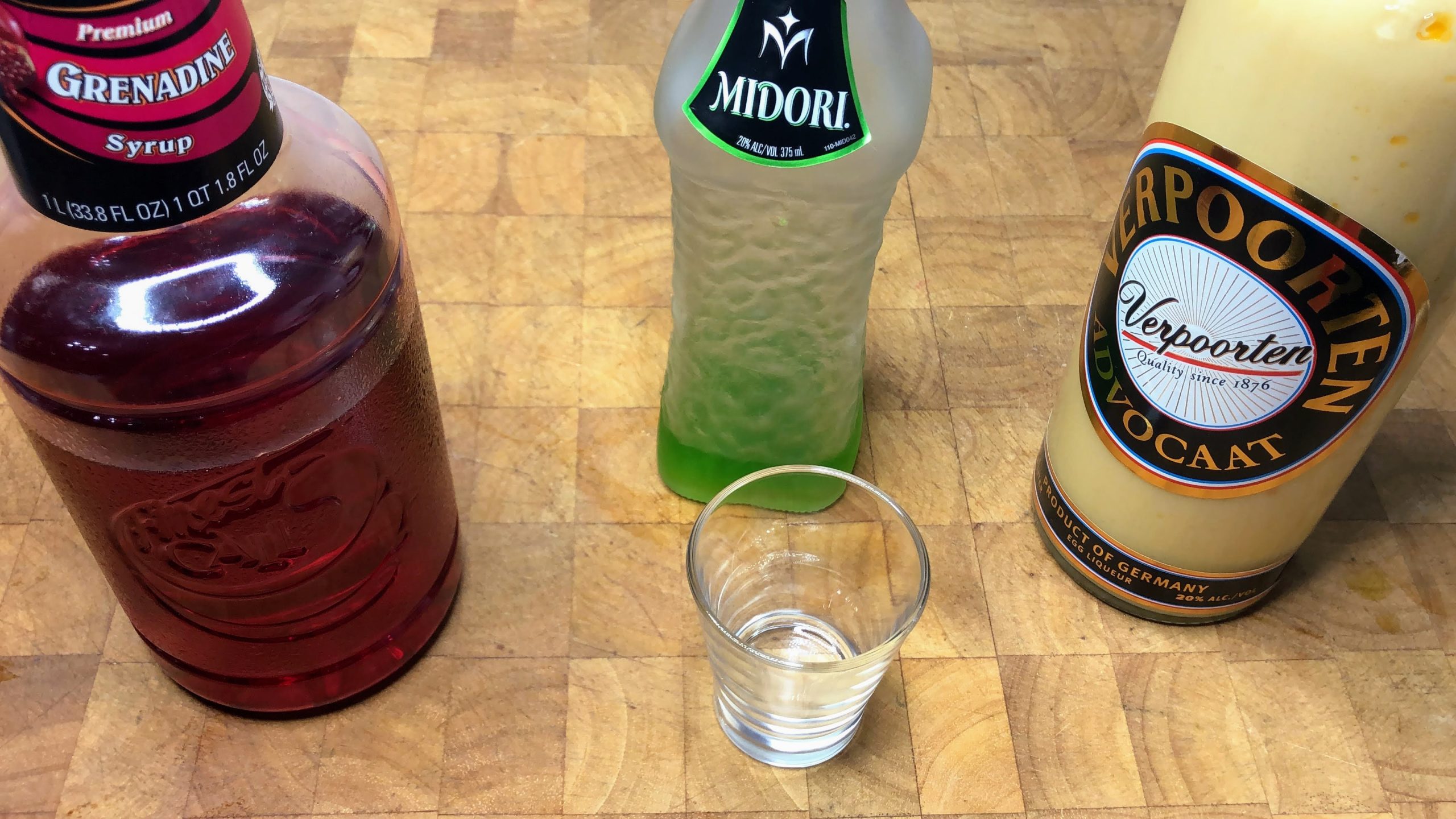 shot glass on wooden table next to bottles of advocaat, midori and grenadine