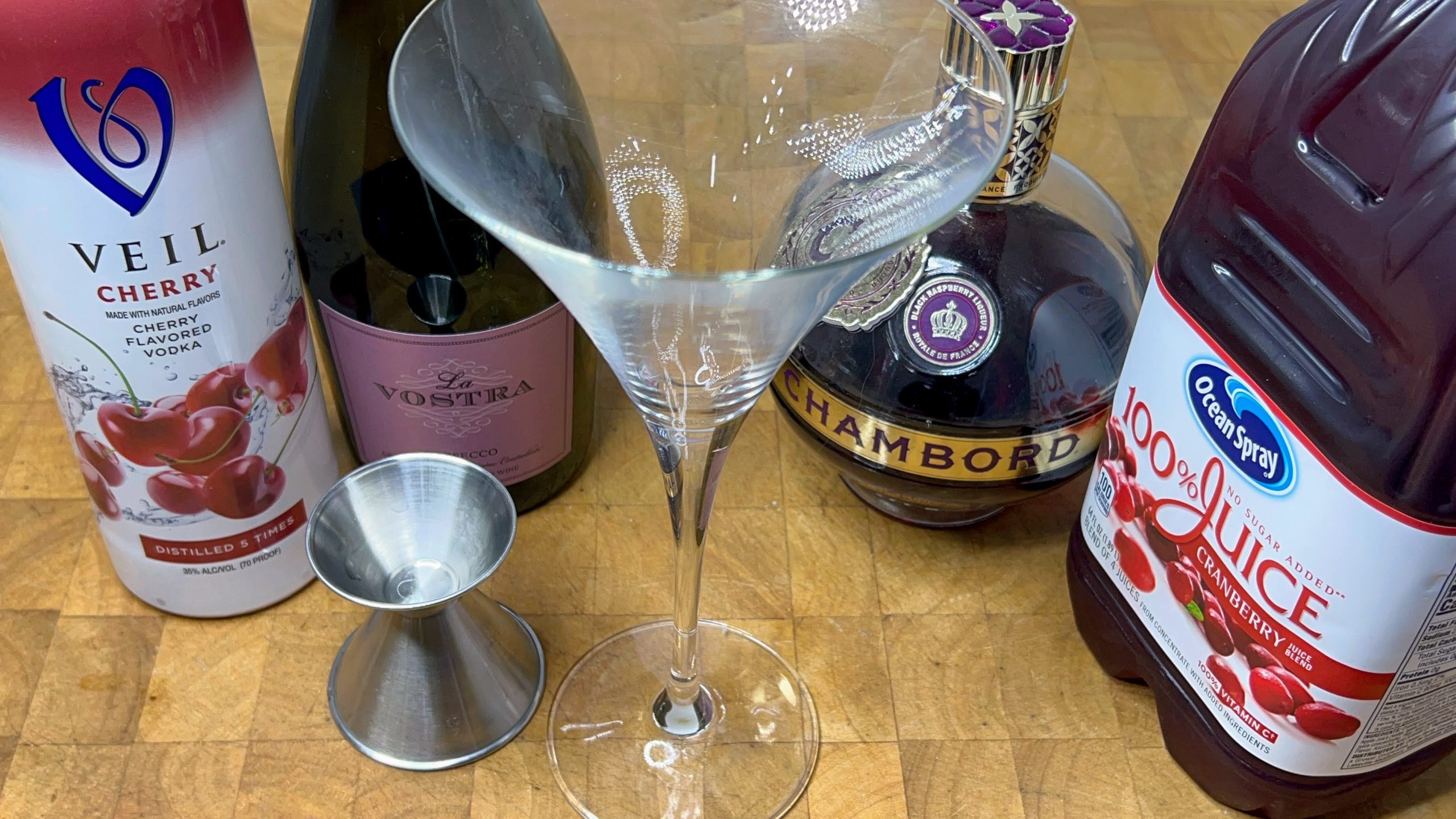 empty martini glass next to jigger, chambord, cranberry juice, cherry vodka and prosecco