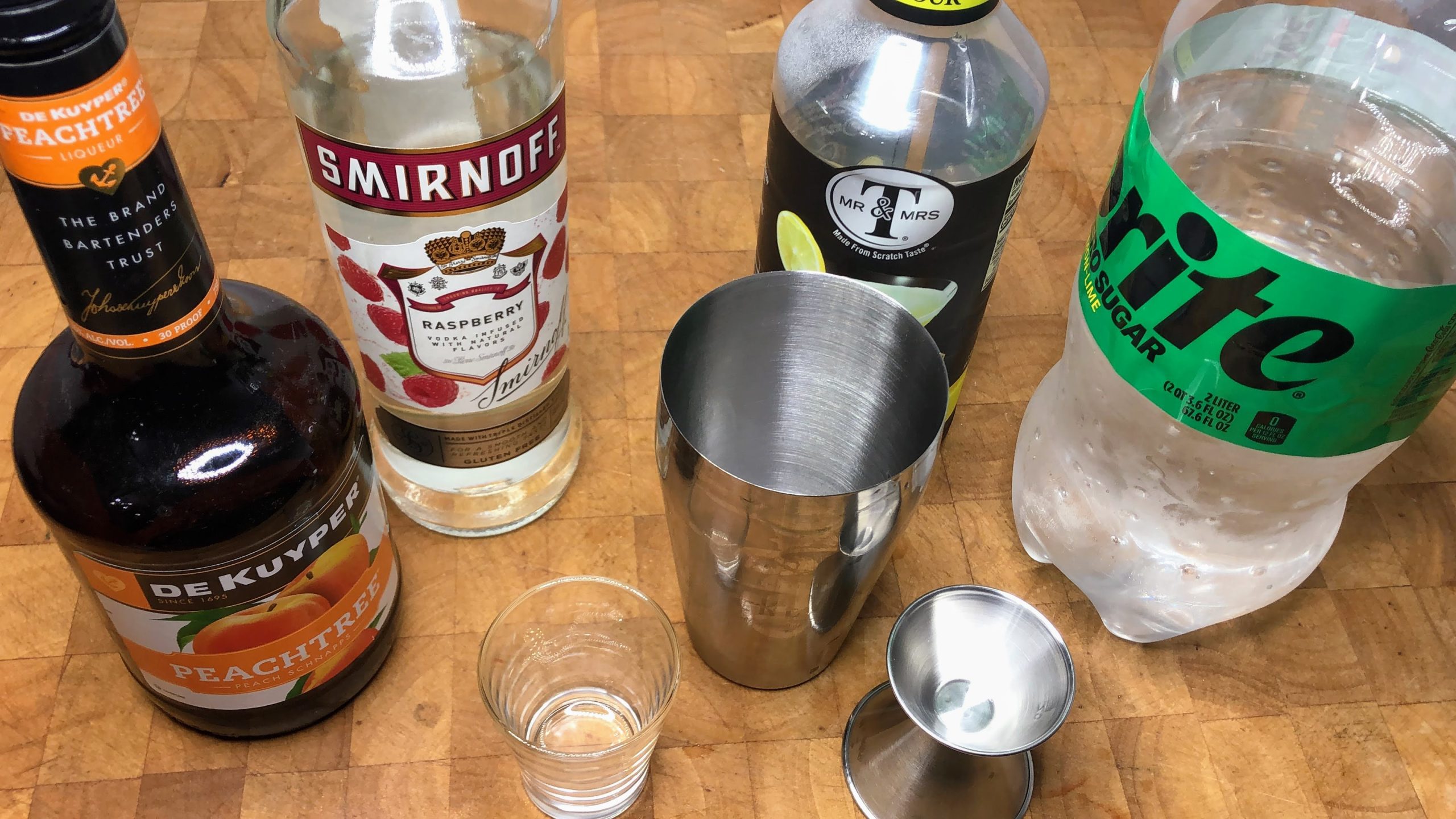 shot glass next to jigger, shaker, bottles of sprite, sour mix, raspberry vodka and peach schnapps