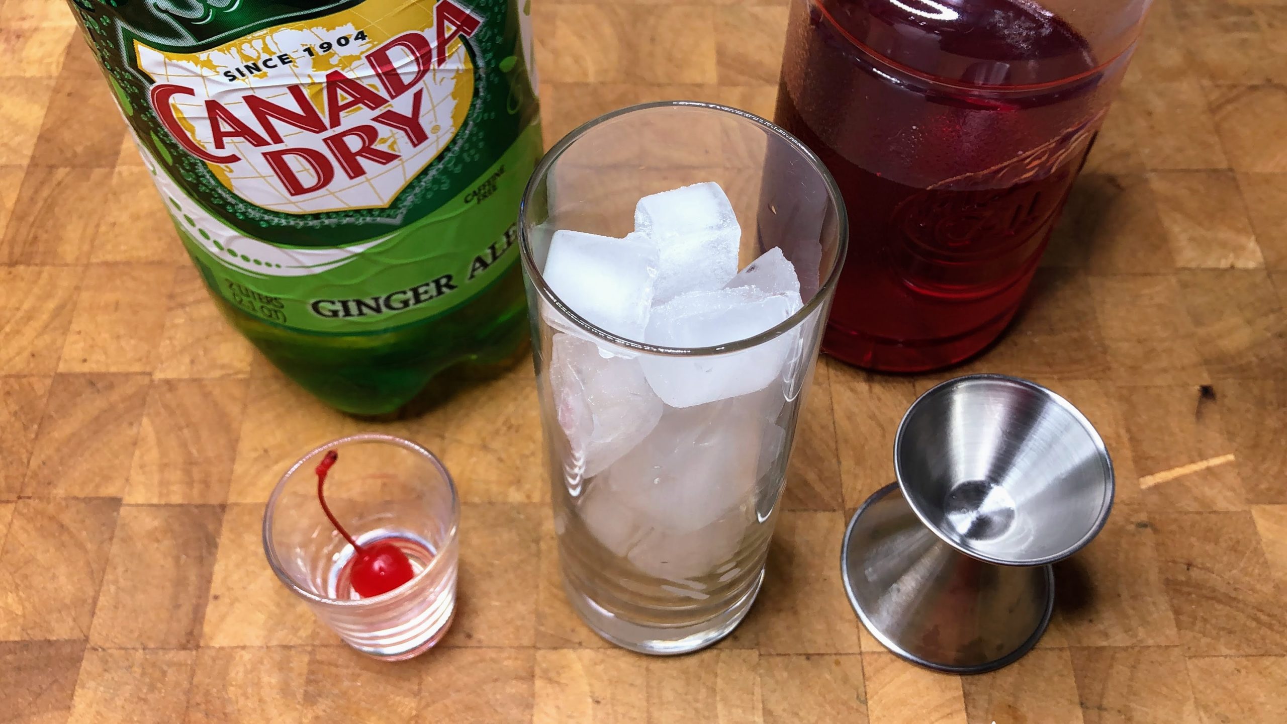 Highball glass filled with ice next to jigger, shot glass with a maraschino cherry and bottles of ginger ale and grenadine.