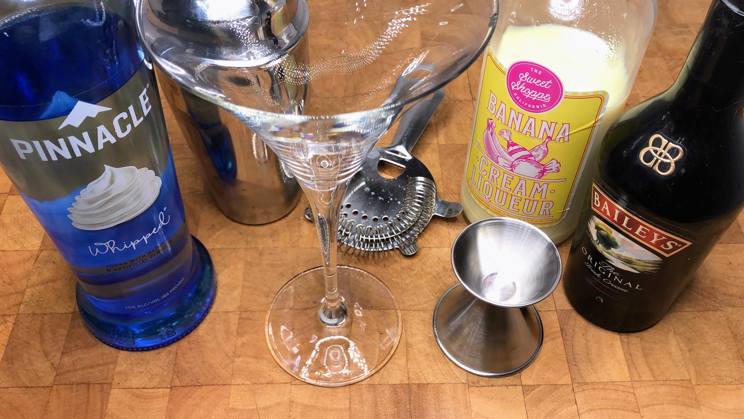 empty martini glass next to jigger, irish cream, banana liqueur, whipped cream vodka and a cocktail shaker