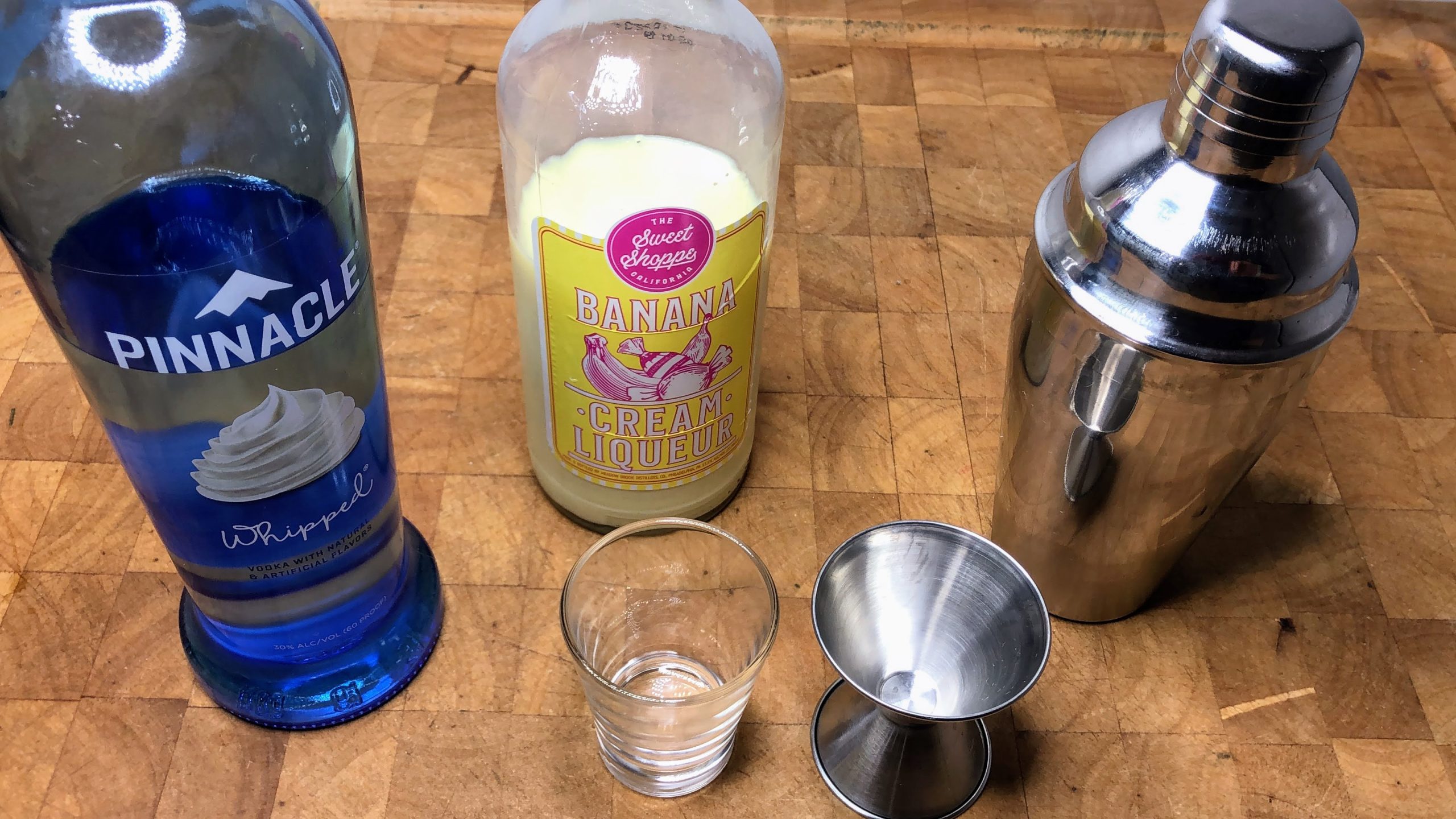 Bottles of whipped cream vodka and banana liqueur next to cocktail shaker, shot glass and jigger.