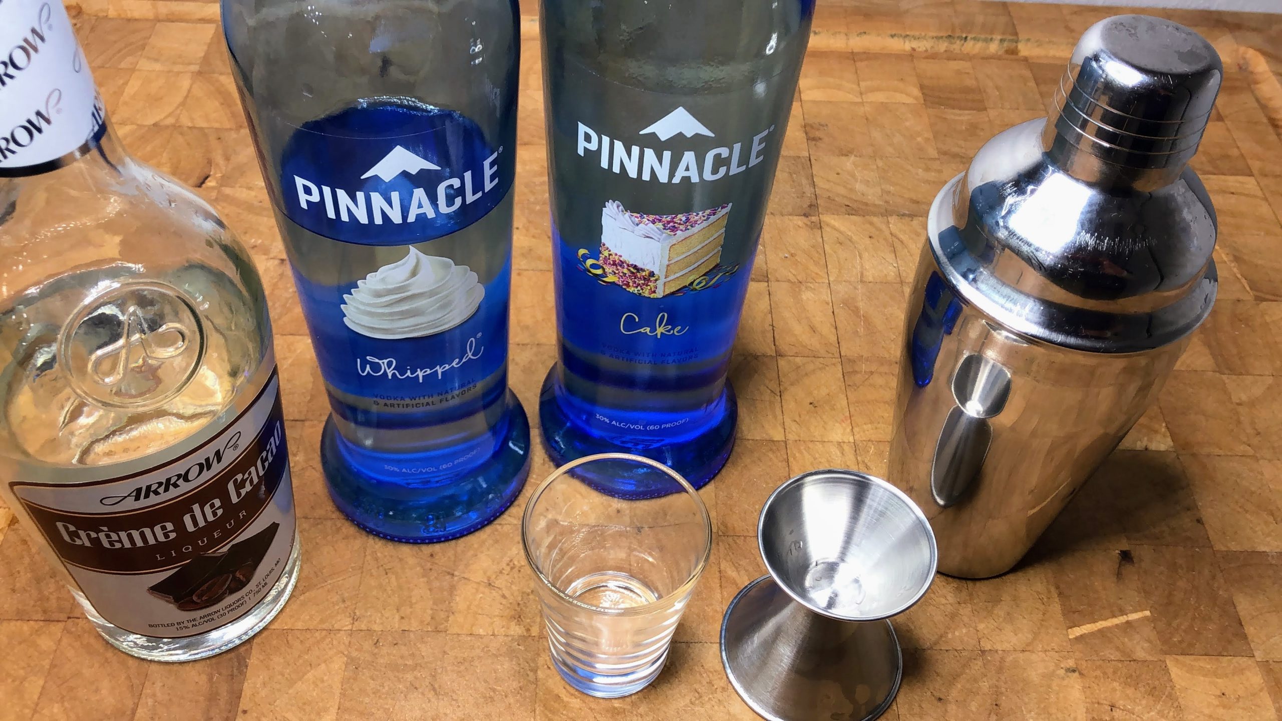 Bottles of creme de cacao, whipped cream vodka and cake vodka next to a shot glass, jigger and cocktail shaker.