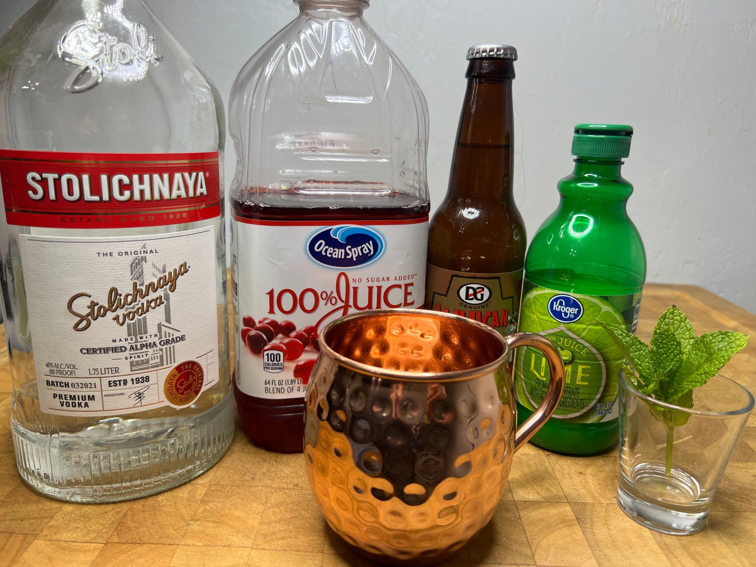 Bottles of vodka, cranberry juice, ginger beer, lime juice next to a copper mug and a sprig of mint.