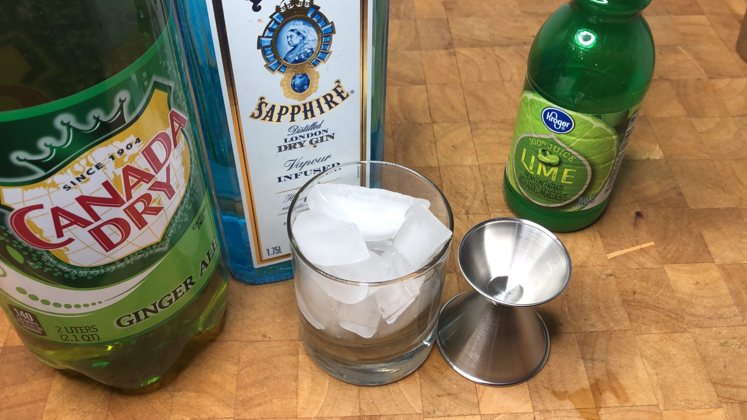 Rocks glass with ice next to a jigger, lime juice, gin and ginger ale.