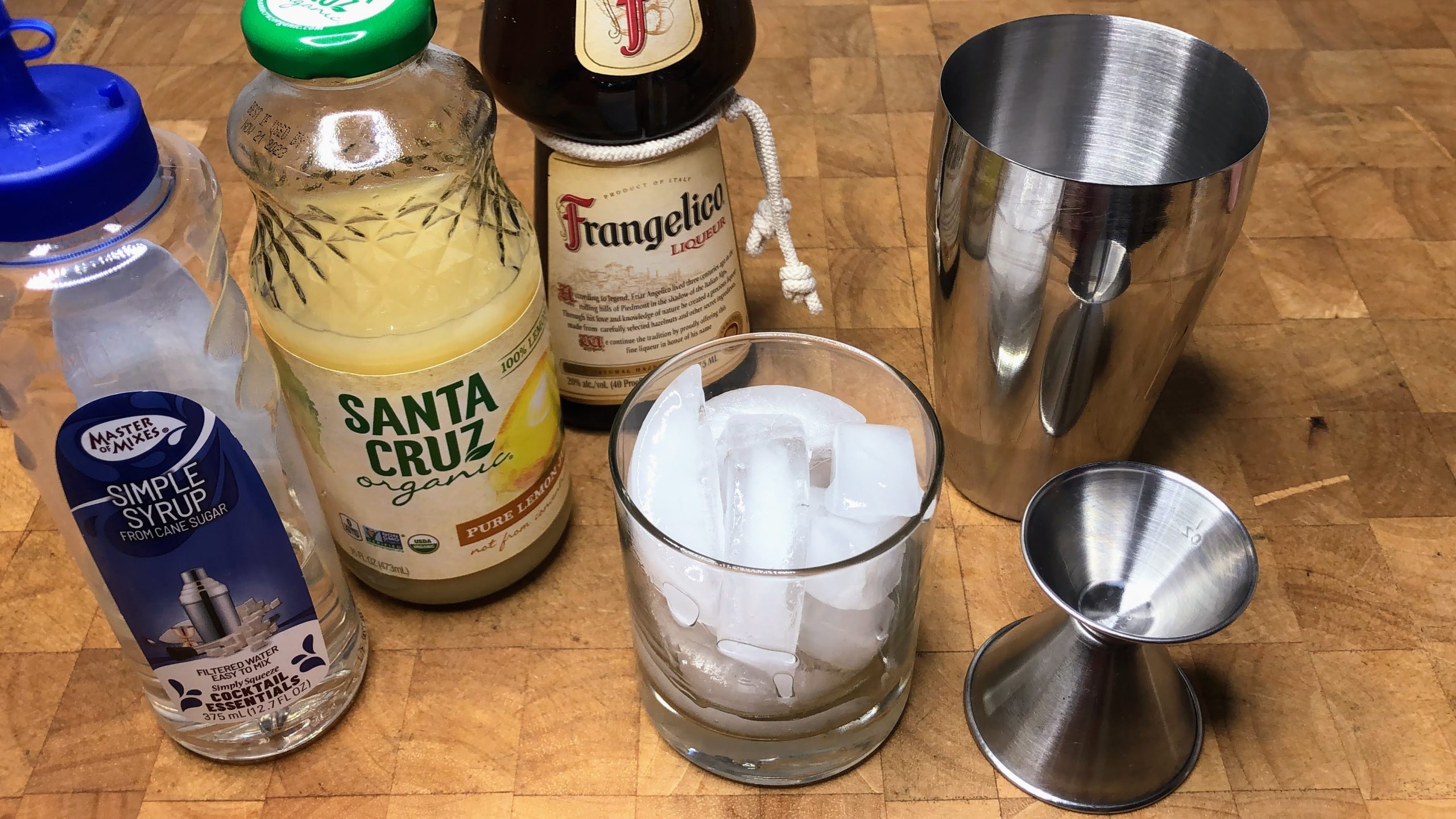 Bottles of simple syrup, lemon juice, frangelico next to a rocks glass, jigger and cocktail shaker.