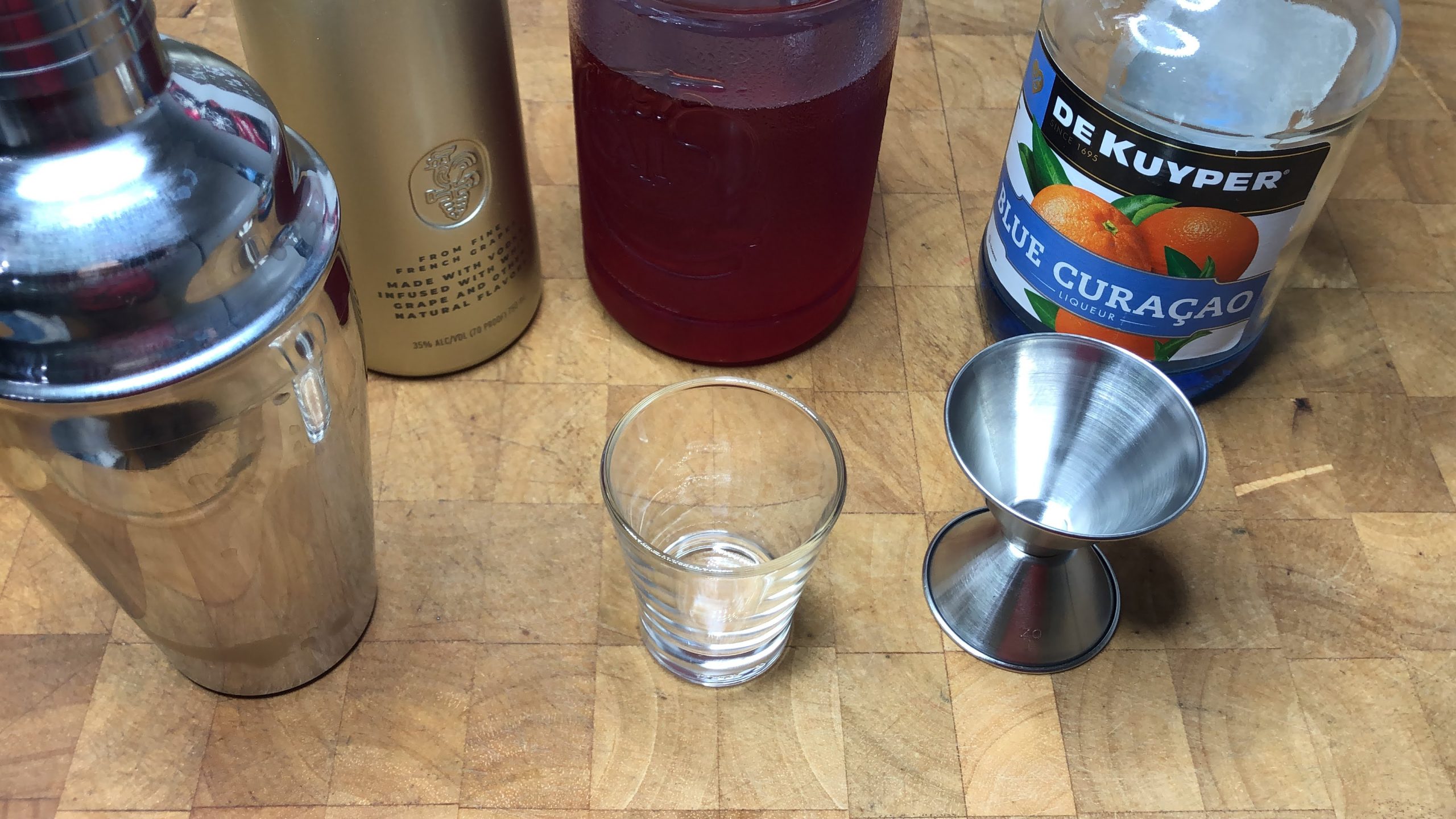 Bottles of blue curacao, grenadine and grape vodka next to shaker, shot glass and jigger.