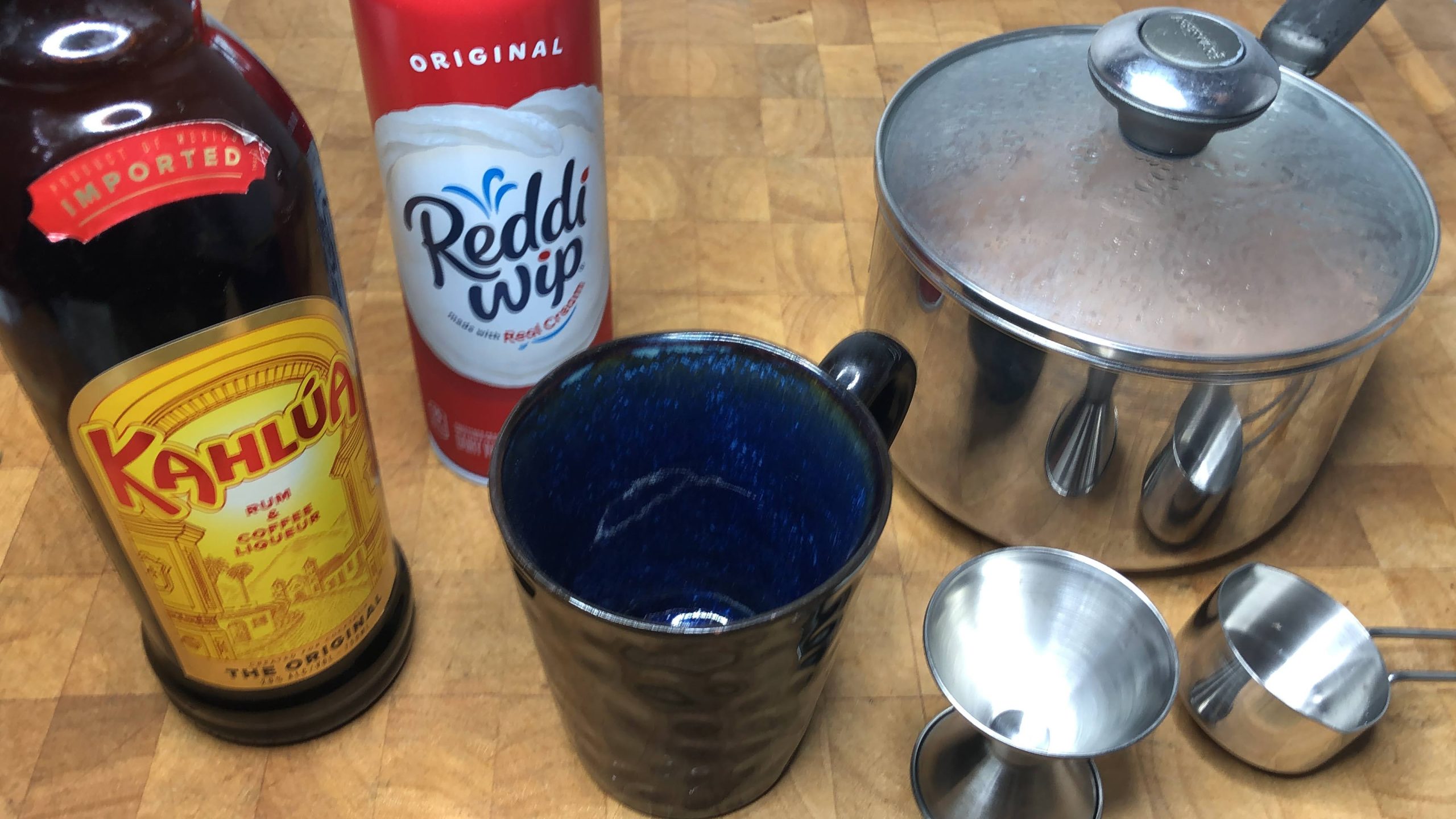 Hot chocolate in a pot next to mug, jigger, measuring cup, whipped cream and kahlua.