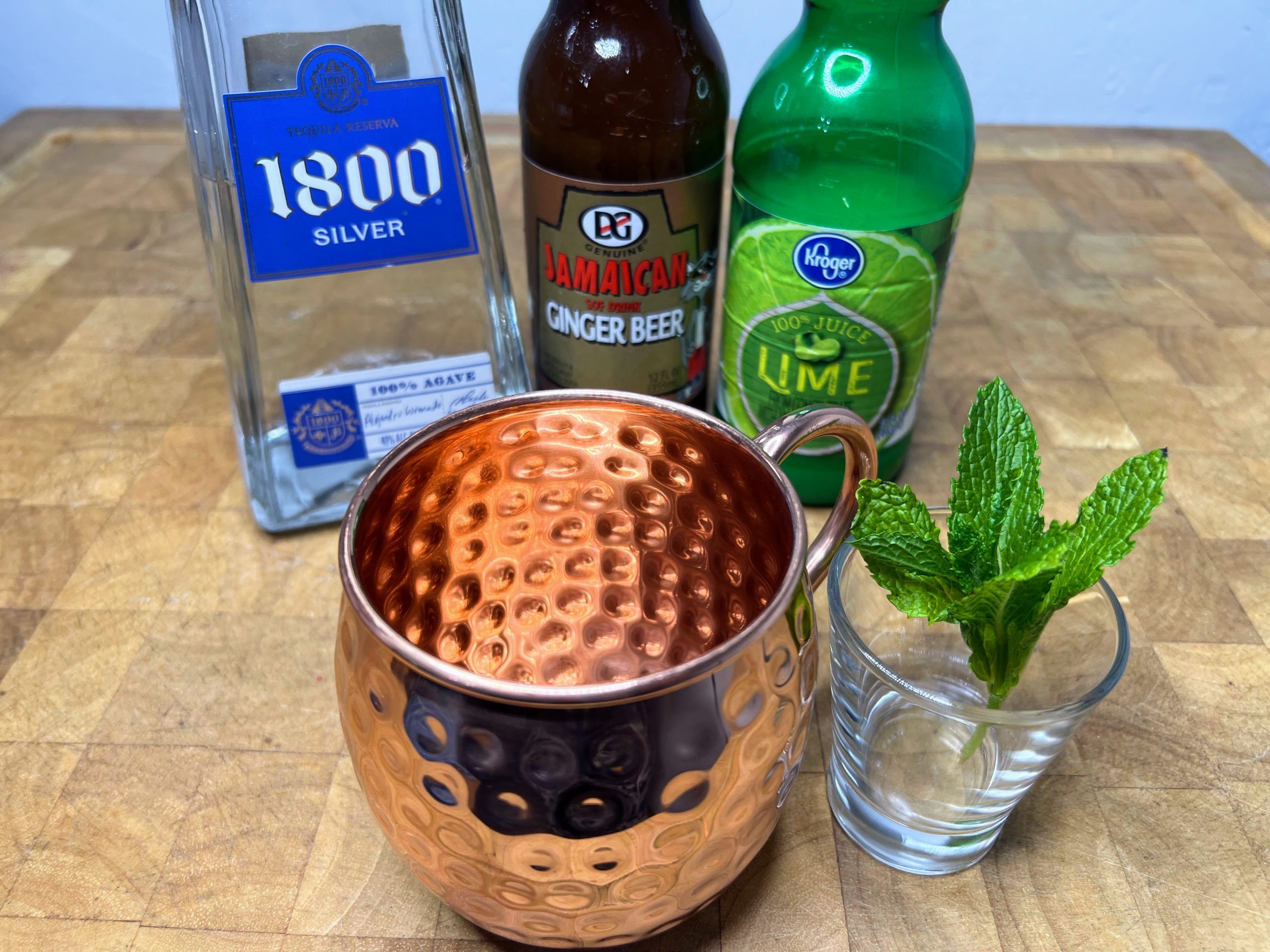 copper mug next to a sprig of mint and bottles of tequila, ginger beer and lime juice