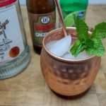 closeup of a moscow mule in a copper mug with mint next to the bottles of ingredients.