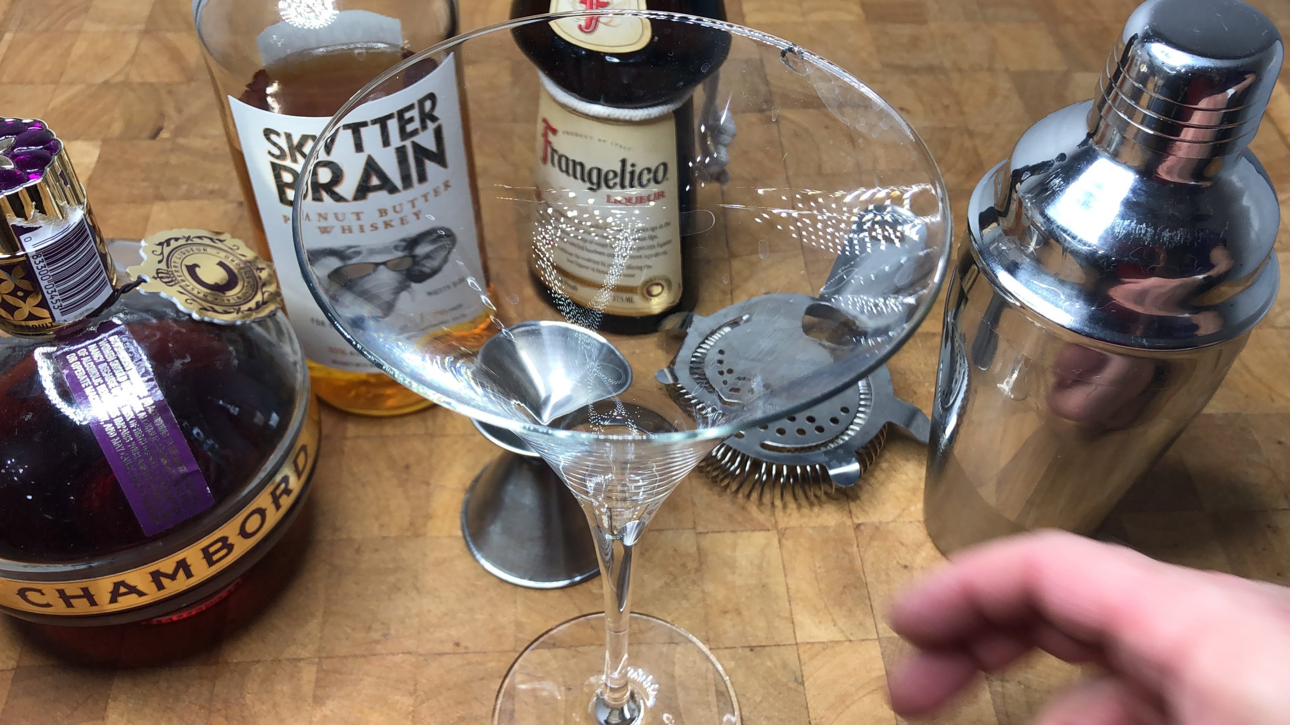 Martini glass next to chambord, peanut butter whiskey, frangelico, jigger, hawthorne strainer and cocktail shaker.