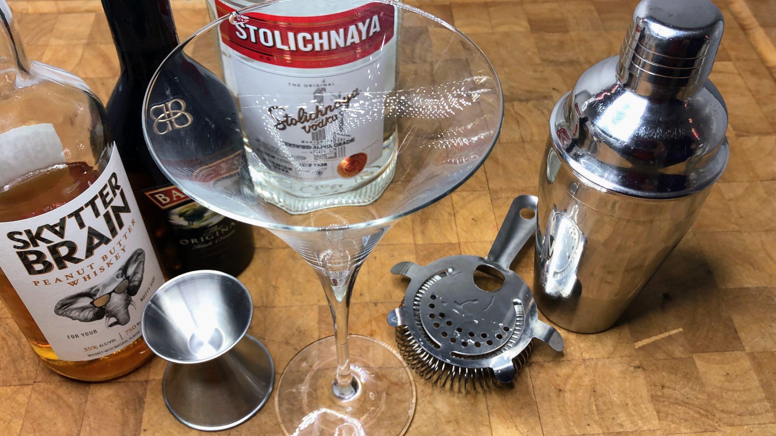 Martini glass next to jigger, hawthorne strainer, cocktail shaker and bottles of peanut butter whiskey, irish cream and vodka.