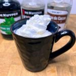 Closeup of a peppermint patty drink in a mug topped with whipped cream and ingredient bottles behind the mug.