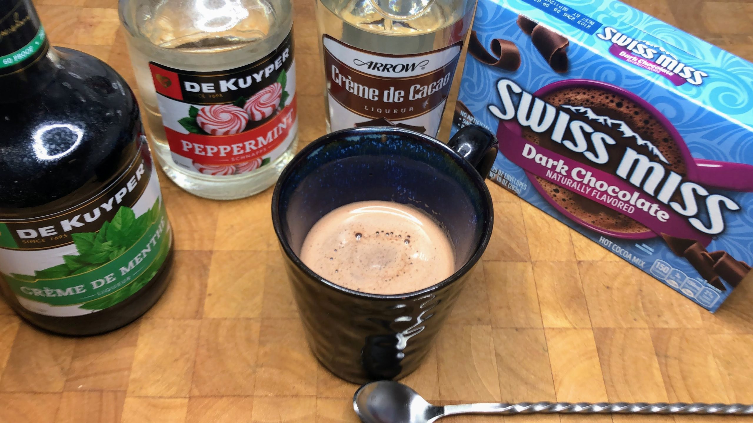 Mug of hot chocolate next to a bar spoon, peppermint schnapps, creme de menthe and white creme de cacao.