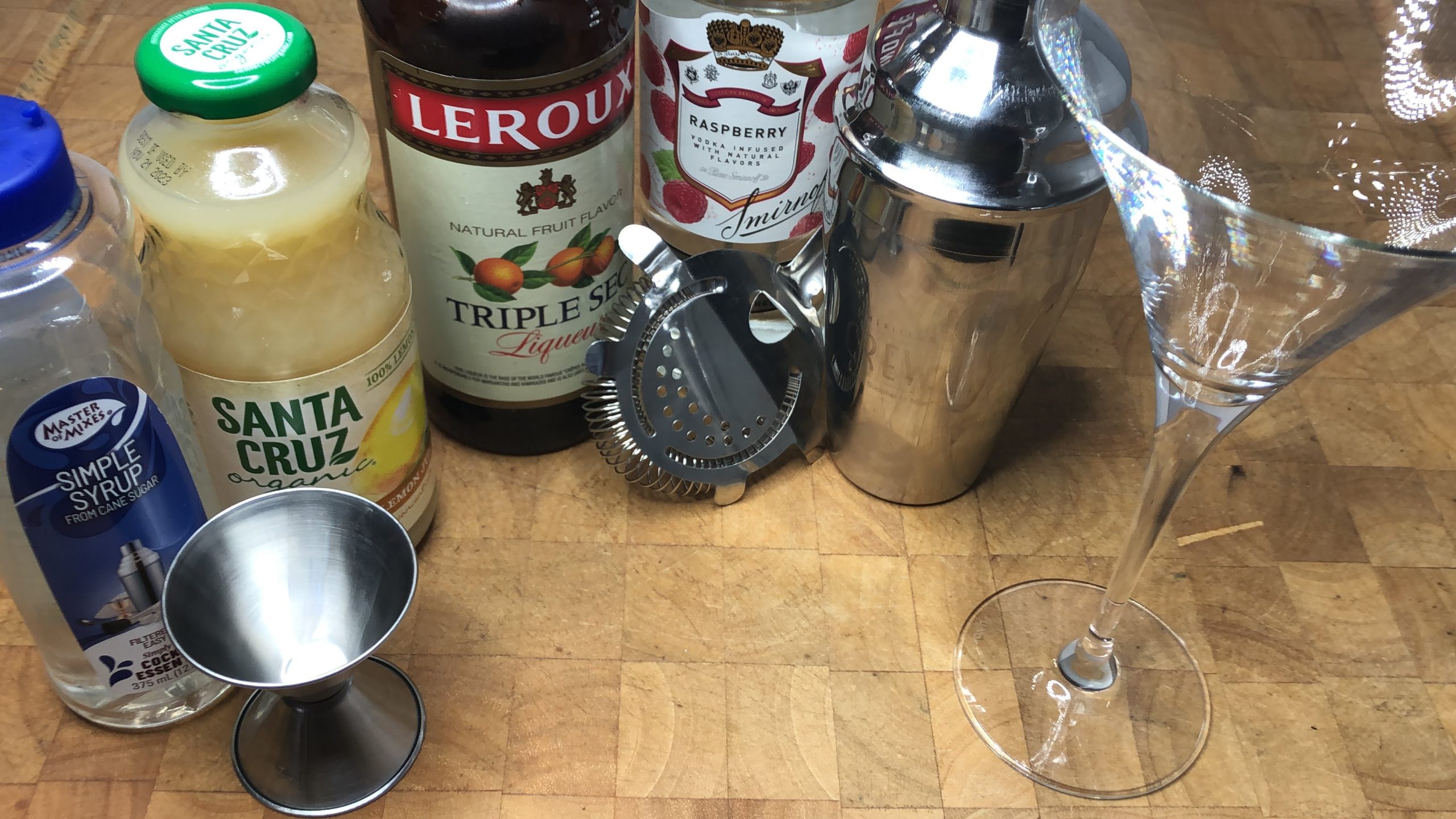 Empty martini glass next to shaker, strainer, jigger, raspberry vodka, triple sec, lemon juice and simple syrup.