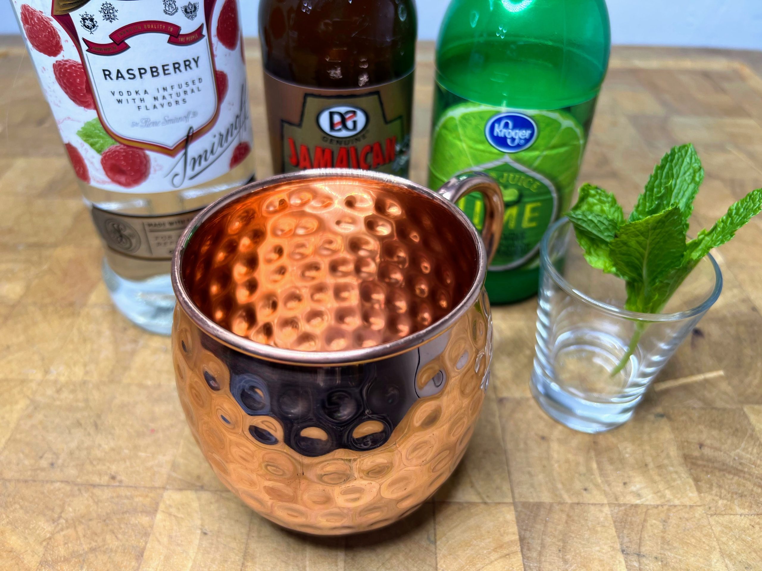 Sprig of mint in a shot glass next to a copper mug and bottles of raspberry vodka, ginger beer and lime juice.