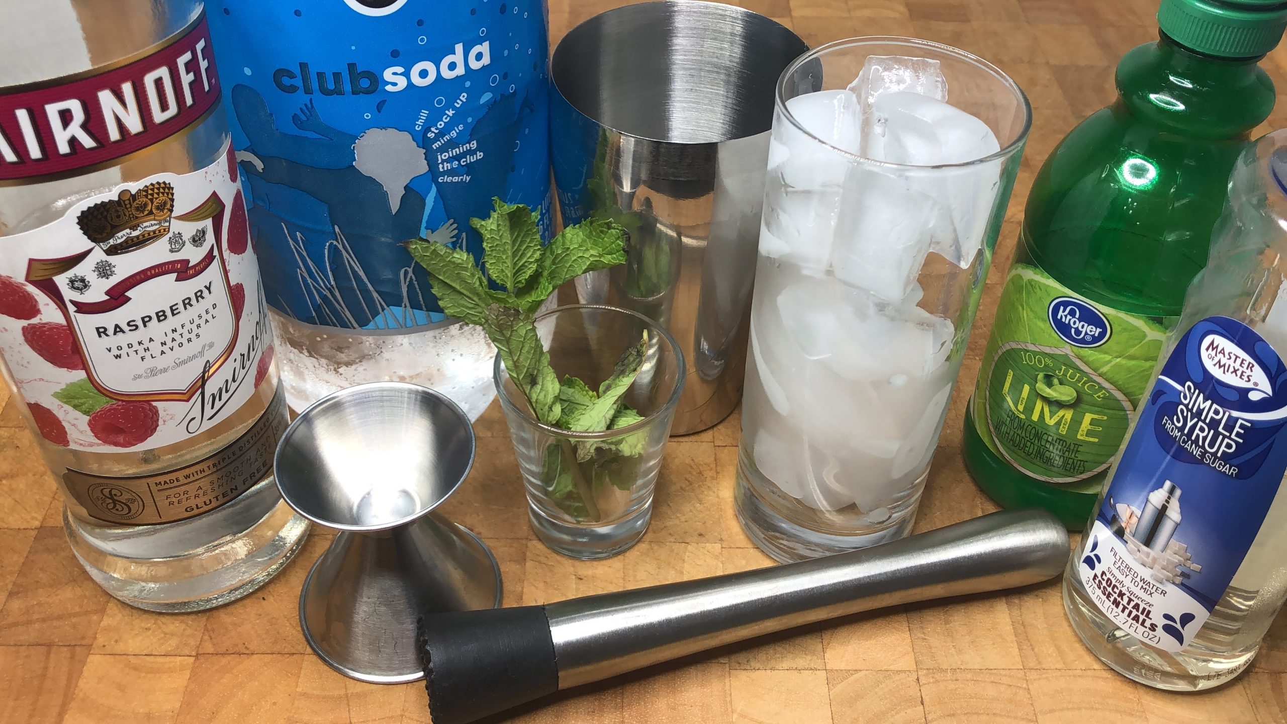 Highball glass with ice next to muddler, jigger, mint, lime juice, simple syrup, shaker, club soda and raspberry vodka.