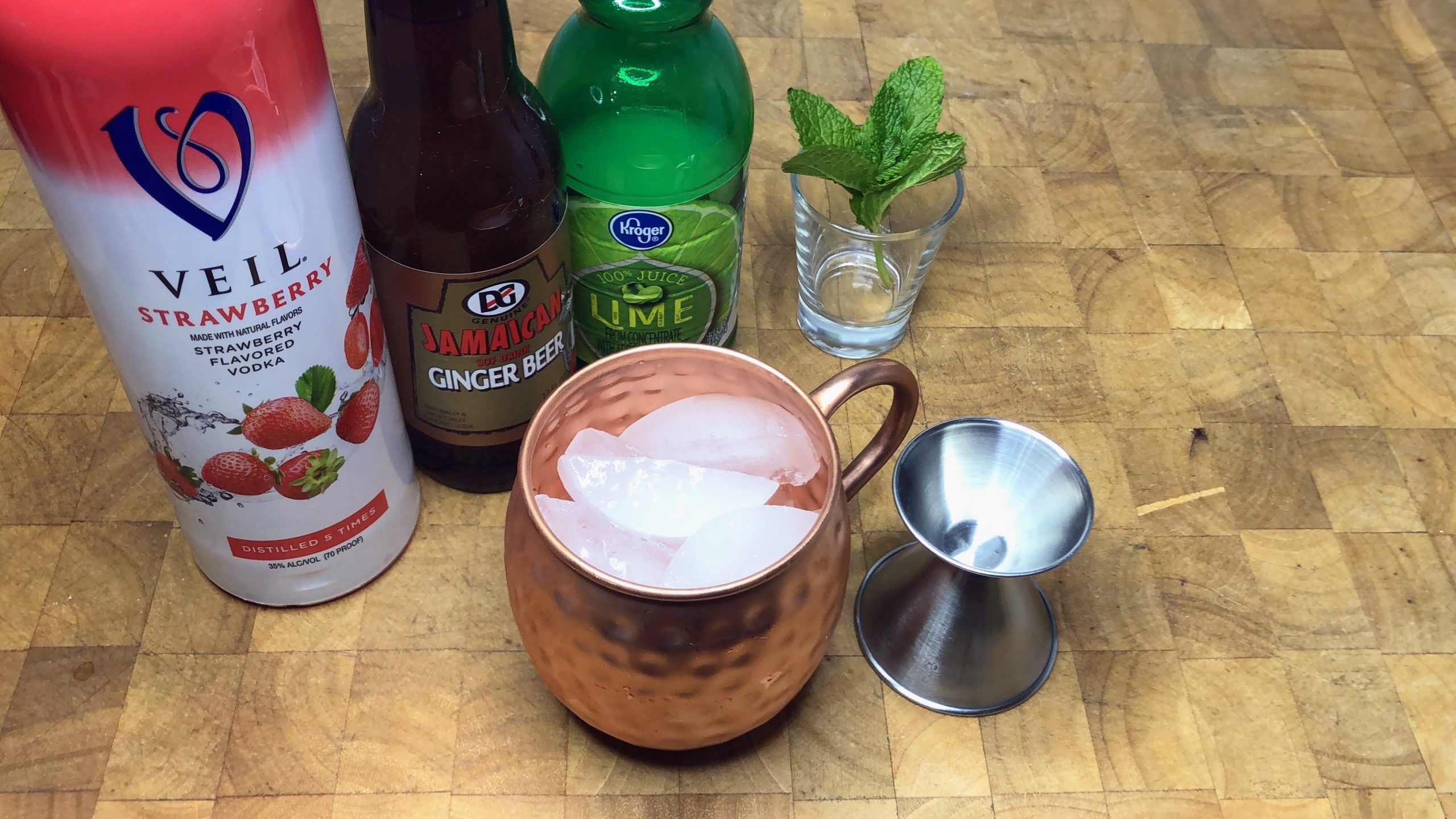 copper mug with ice next to jigger, sprig of mint, strawberry vodka, ginger beer and lime juice.